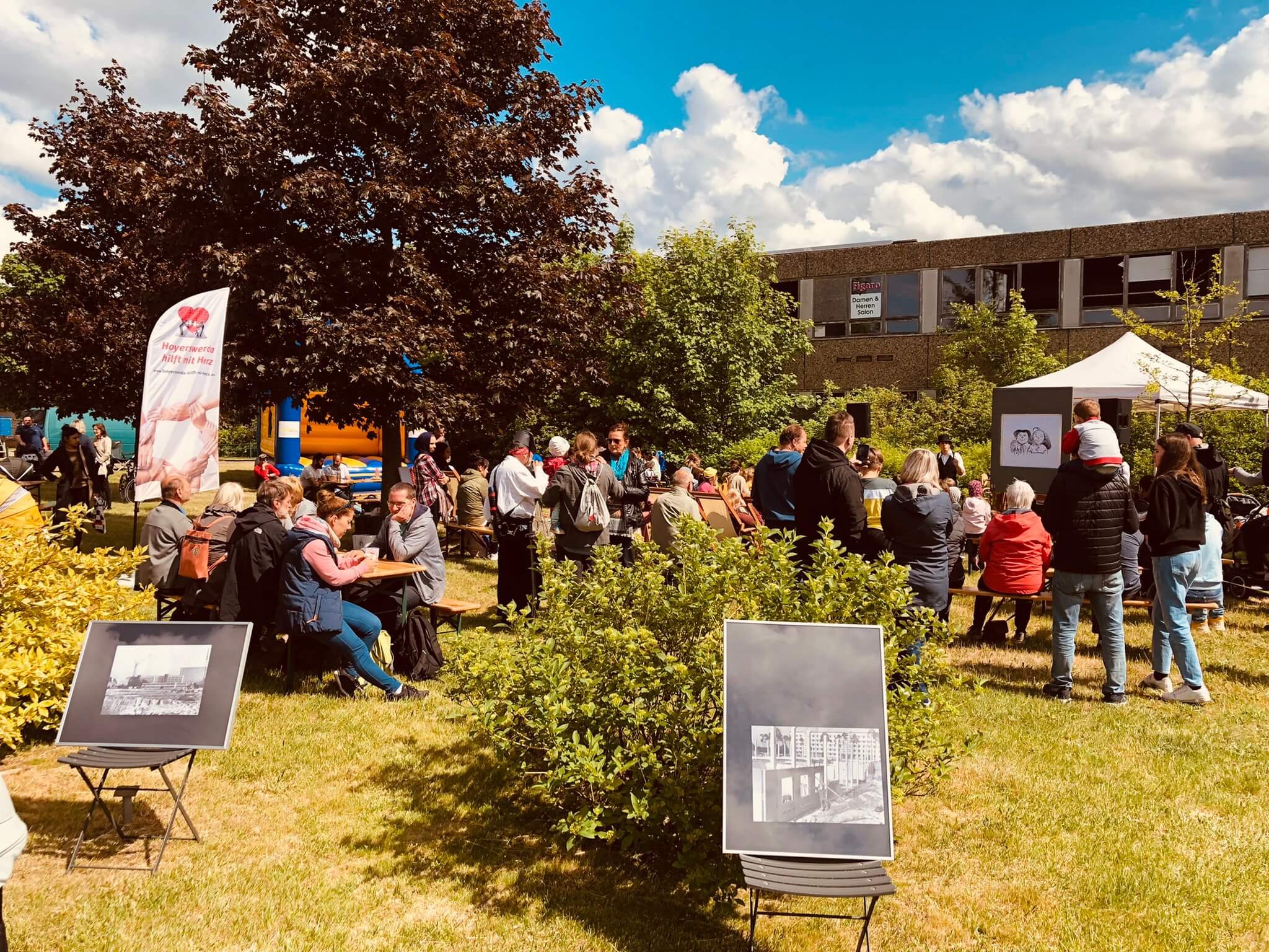 Menschen besuchen ein Sommerfest im Park mit Foto-Aufstellern, Sitzbänken und einer Hüpfburg.