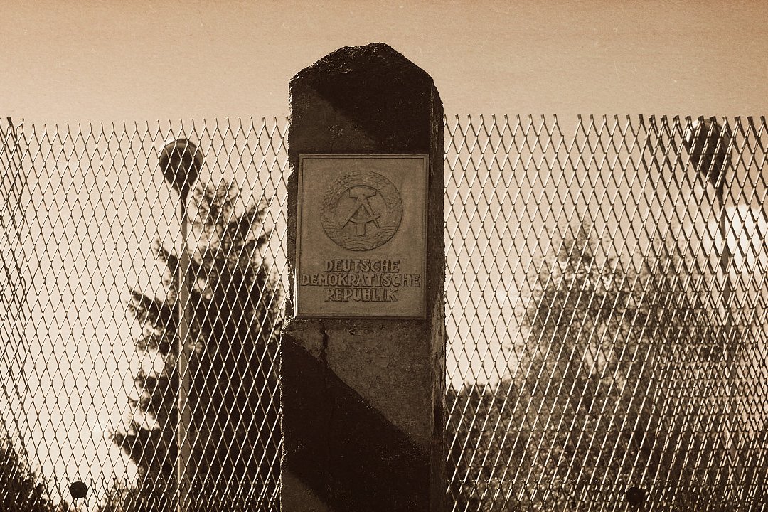 Sepiafarbenes Foto eines Grenzzauns aus Metallgitter mit einem massiven Betonpfosten in der Mitte. Auf dem Pfosten ist eine Tafel mit dem Emblem und der Aufschrift „Deutsche Demokratische Republik“ angebracht. Hinter dem Zaun sind Bäume und ein runder Grenzpfosten zu erkennen.