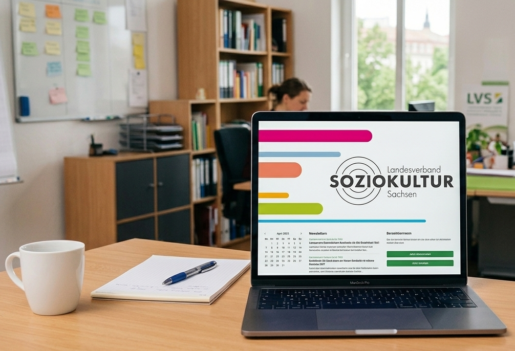 Laptop auf Schreibtisch im Büro, zeigt Website „Soziokultur Sachsen“, Personen arbeiten im Hintergrund
