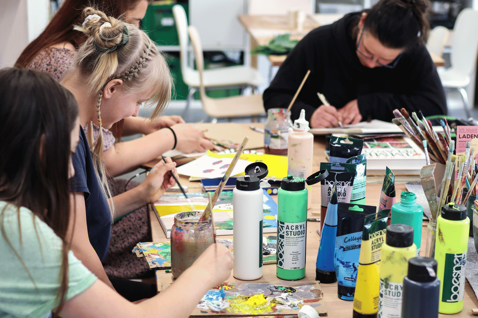 Eine Nahaufnahme einer kreativen Arbeitssituation an einem langen Holztisch. Im Vordergrund malen mehrere junge Menschen konzentriert mit Pinseln auf Papier und Pappe. Auf dem Tisch verteilt stehen zahlreiche Utensilien: bunte Acrylfarben in Tuben und Flaschen der Marke Boesner, Gläser mit Malwasser und ein großer Behälter voller Pinsel. Im Hintergrund ist eine weitere Person zu sehen, die über ihre Arbeit gebeugt ist. Die Szene vermittelt eine Atmosphäre von künstlerischem Schaffen und Fokus.