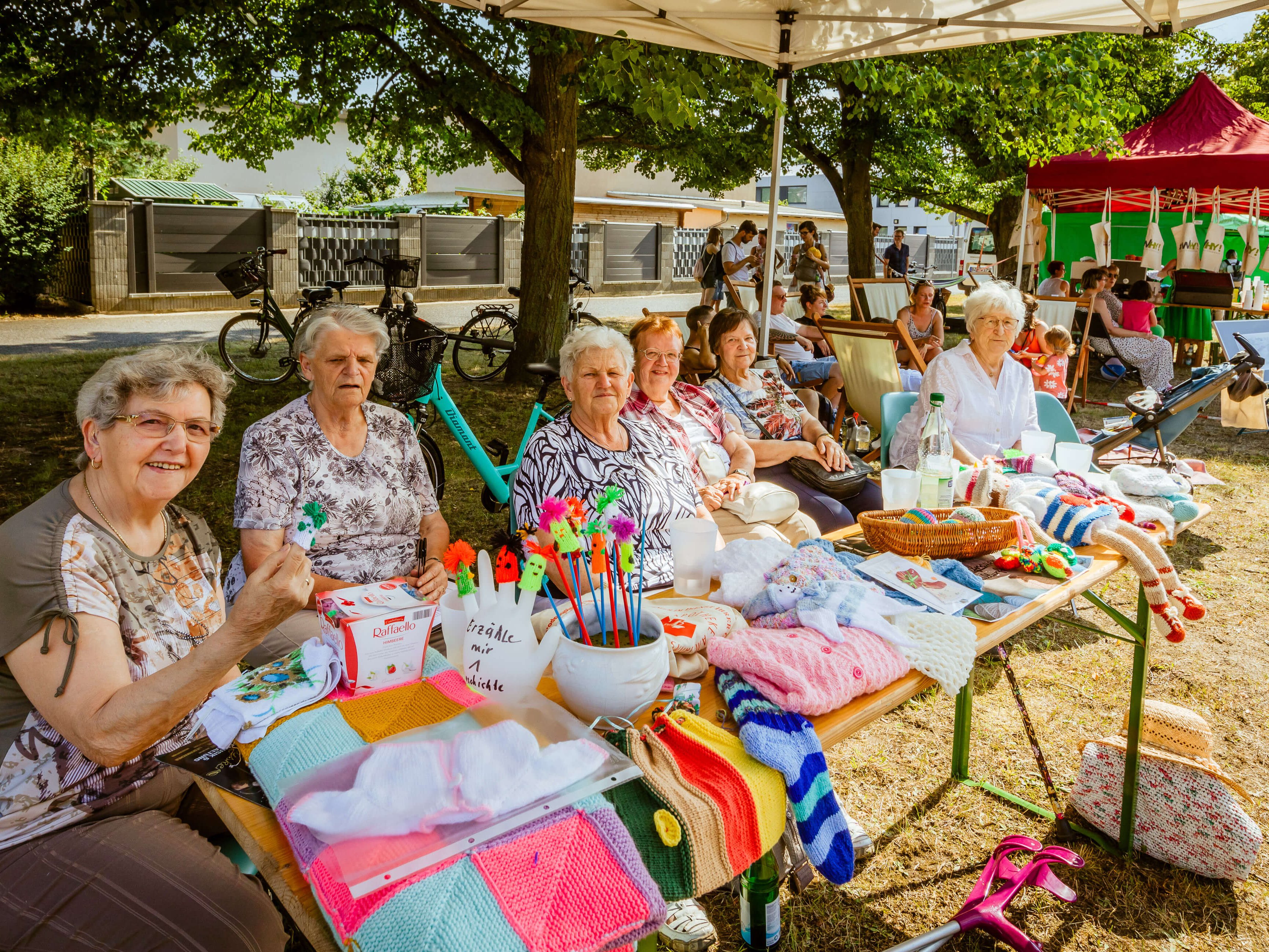 Seniorinnen an einem Stand mit bunter Strickware auf einem sonnigen Nachbarschaftsfest.