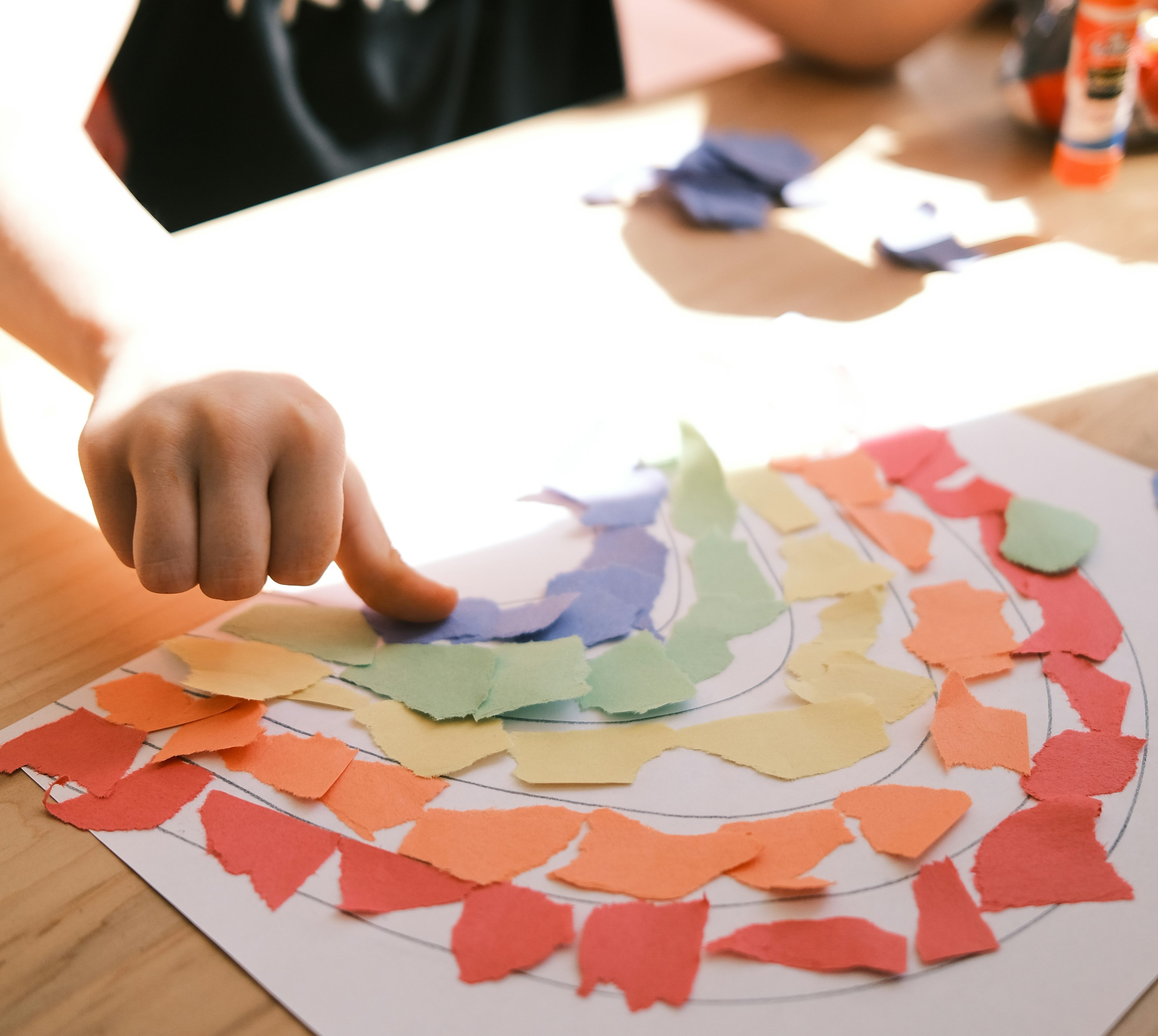 Ein Kind legt bunte, zerrissene Papierstücke auf ein großes Blatt Papier und gestaltet daraus ein kreisförmiges, regenbogenähnliches Muster an einem Tisch.