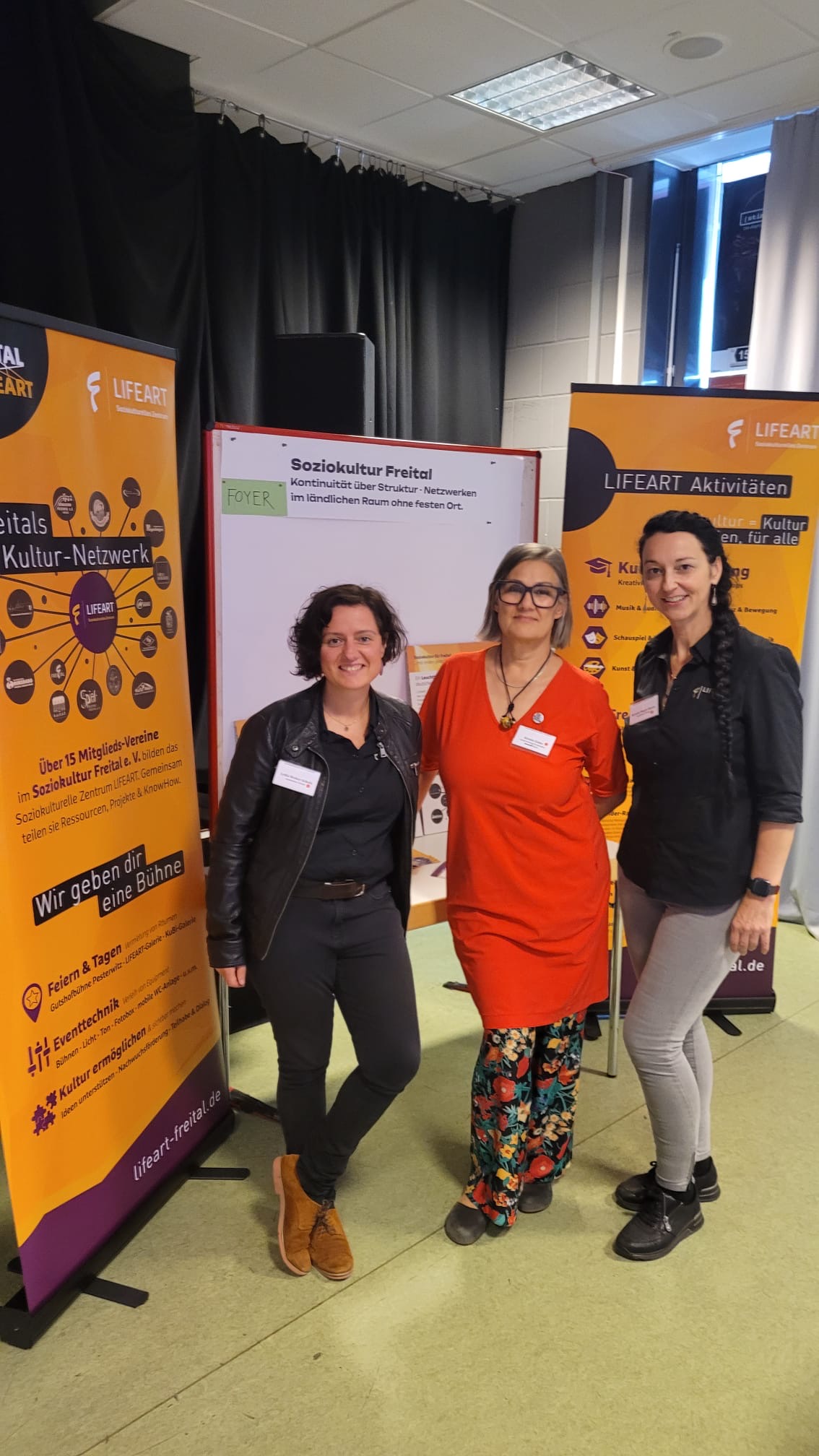 Drei Frauen stehen bei einer Kulturveranstaltung vor Informations- und Roll-up-Bannern von LIFEART. Im Hintergrund sind ein Präsentationsbildschirm mit dem Titel „Soziokultur Freital“ sowie weitere Veranstaltungsmaterialien zu sehen.