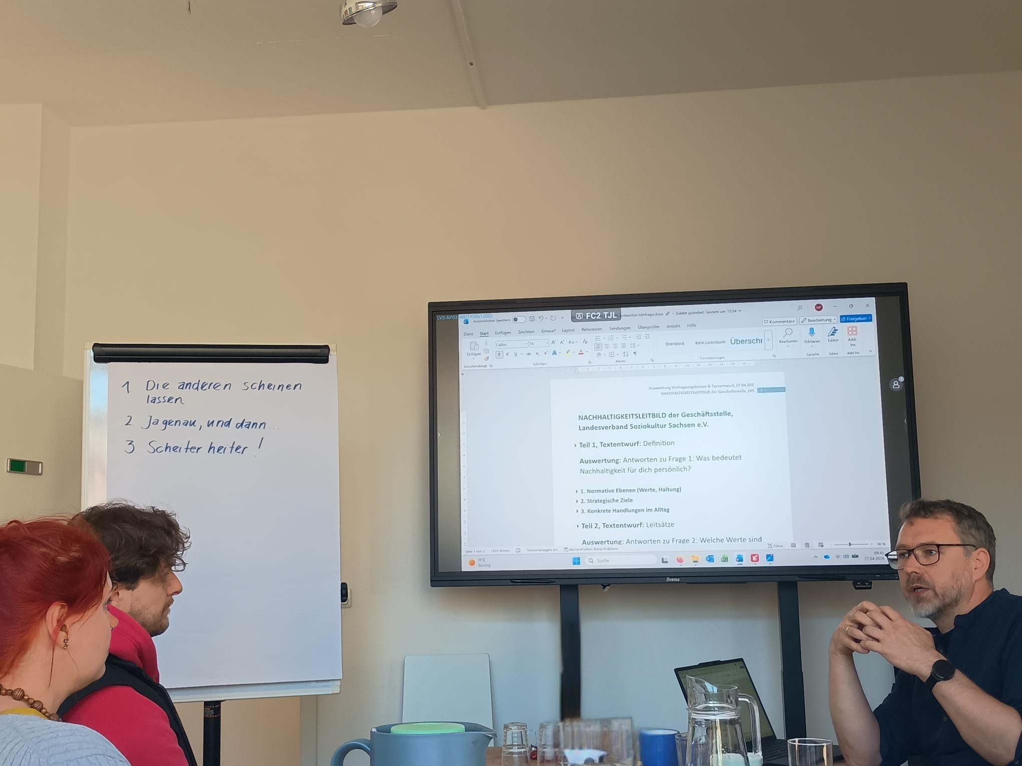 Drei Personen in Workshop, Flipchart mit Stichpunkten, Präsentation auf Bildschirm.