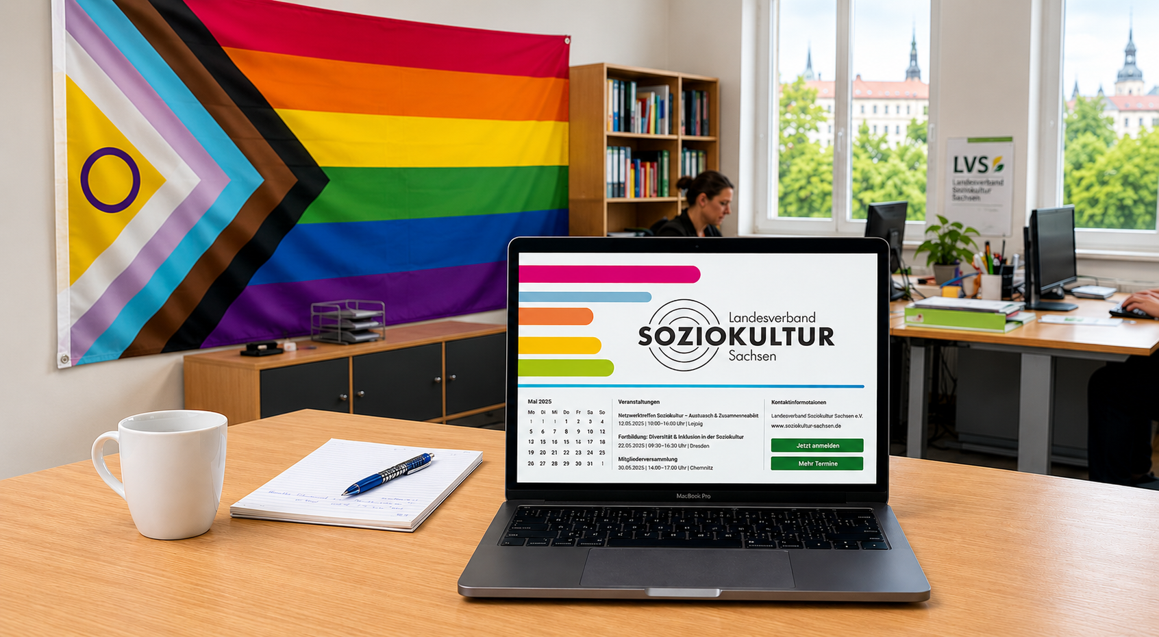 Laptop auf einem Schreibtisch vor einer Progress-Pride-Flagge im Hintergrund eines Büros.