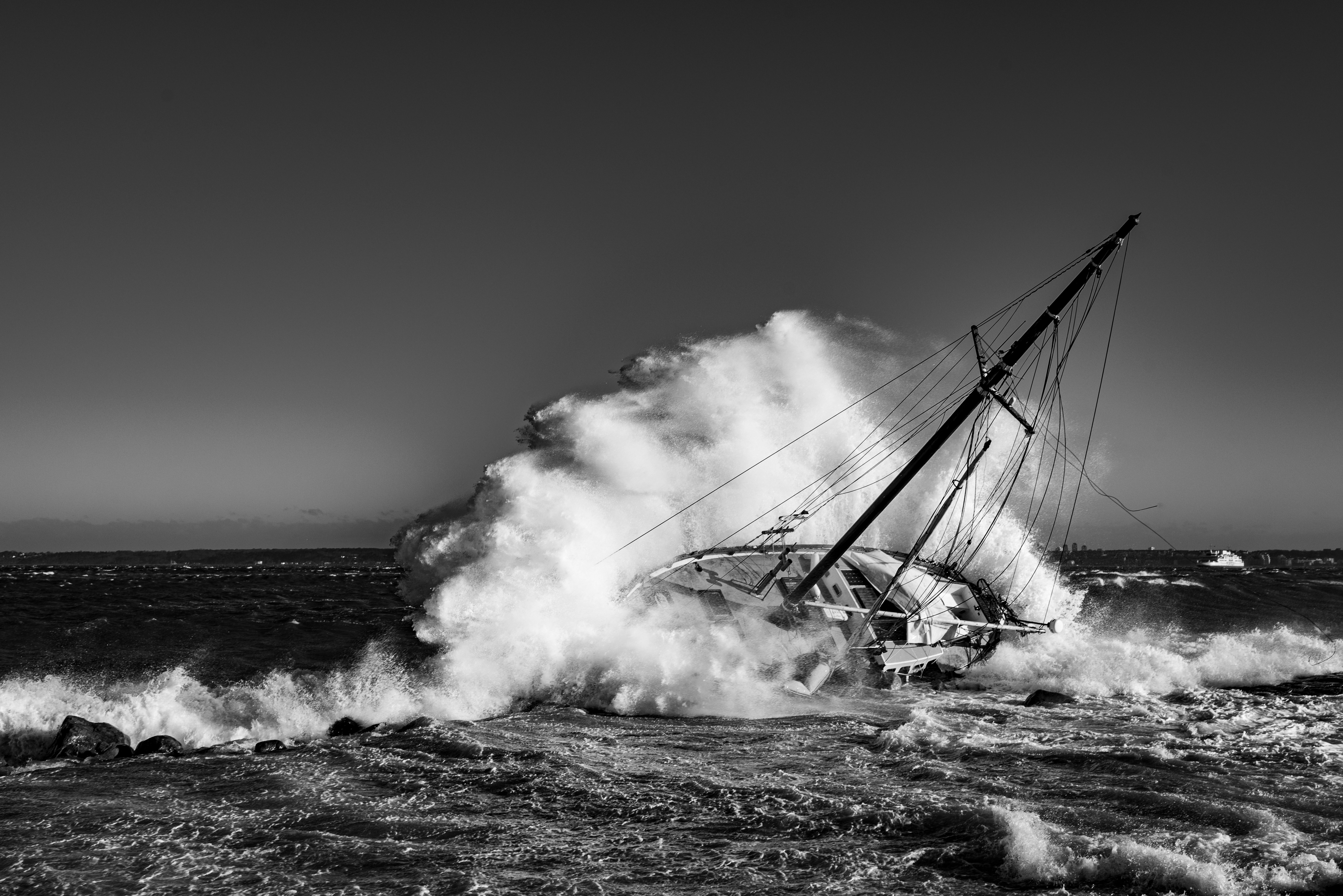 Schwarz-Weiß-Fotografie eines gekenterten Segelboots, das von hohen, schäumenden Wellen getroffen wird. Das Boot liegt stark zur Seite geneigt im stürmischen Meer; Masten, Takelage und Teile des Rumpfs sind sichtbar. Im Hintergrund erstreckt sich ein dunkler Horizont unter einem wolkenverhangenen Himmel.