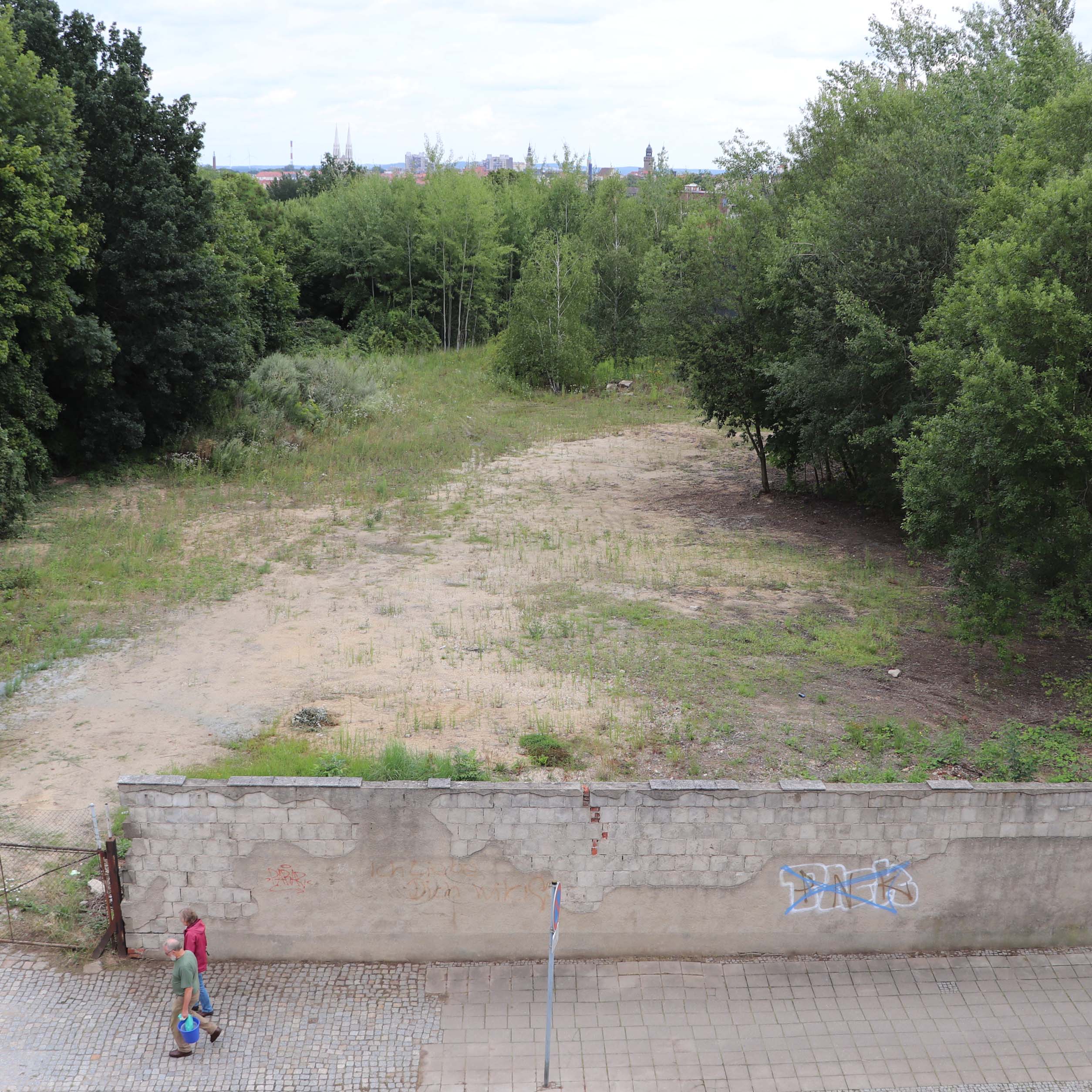 Brachenfläche mit Bäumen, davor Mauer mit Graffiti und zwei Personen auf Gehweg