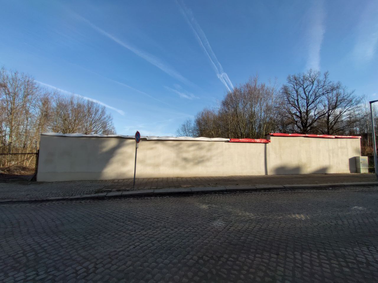 Helle Mauer an Kopfsteinpflasterstraße, kahle Bäume dahinter, blauer Himmel mit Kondensstreifen