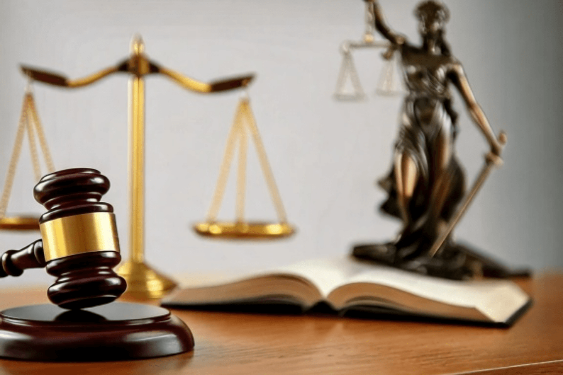Nahaufnahme eines hölzernen Richterhammers mit goldfarbenem Ring auf einem Tisch. Im Hintergrund sind unscharf eine goldene Waage, eine Justitia-Figur mit Schwert und Waage sowie ein aufgeschlagenes Buch zu sehen.