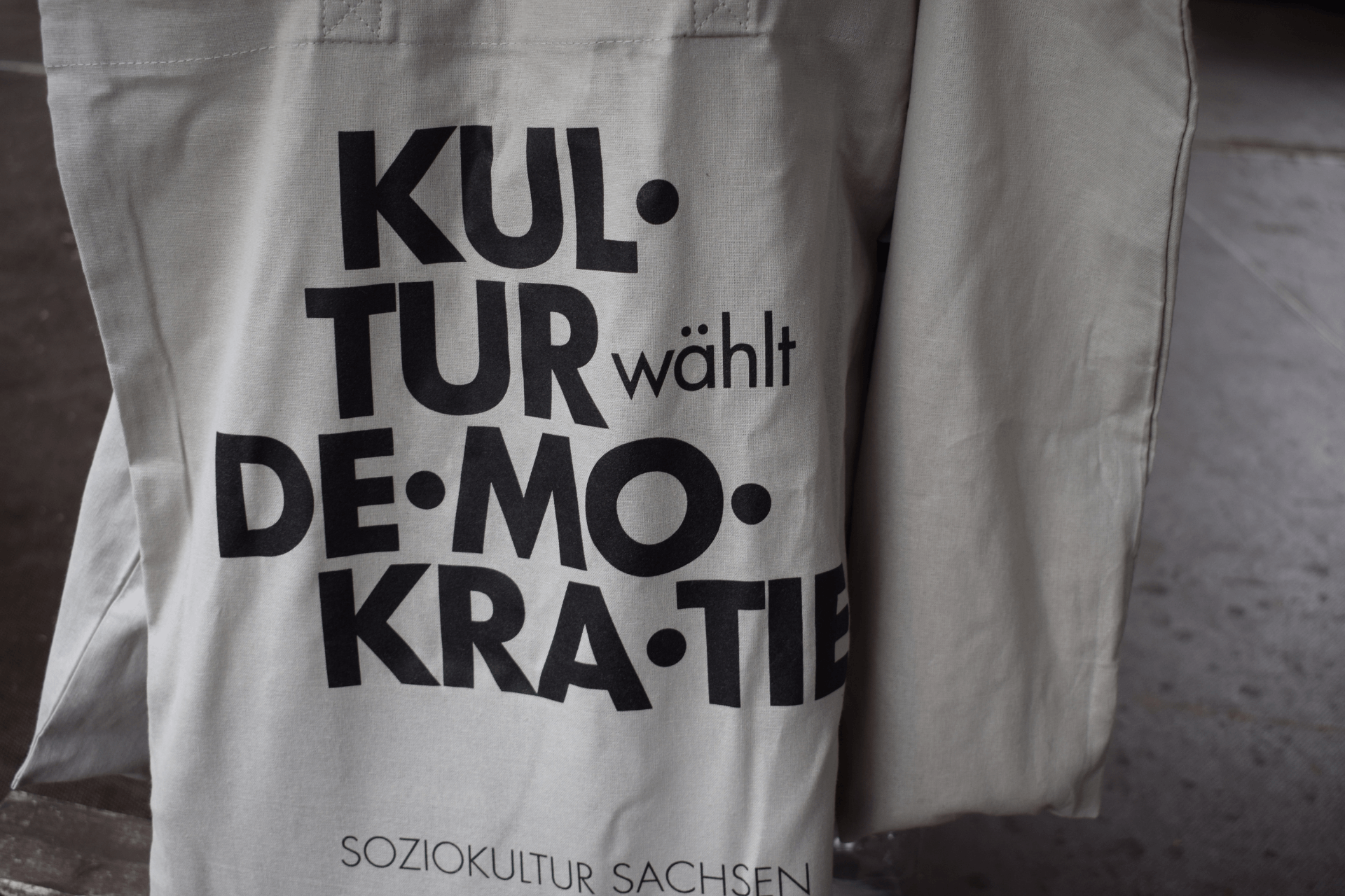 Beiger Stoffbeutel mit schwarzer, groß gedruckter Aufschrift „Kultur wählt Demokratie“; darunter steht kleiner „Soziokultur Sachsen“. Der Beutel hängt vor einem unscharfen Hintergrund im Außenbereich.