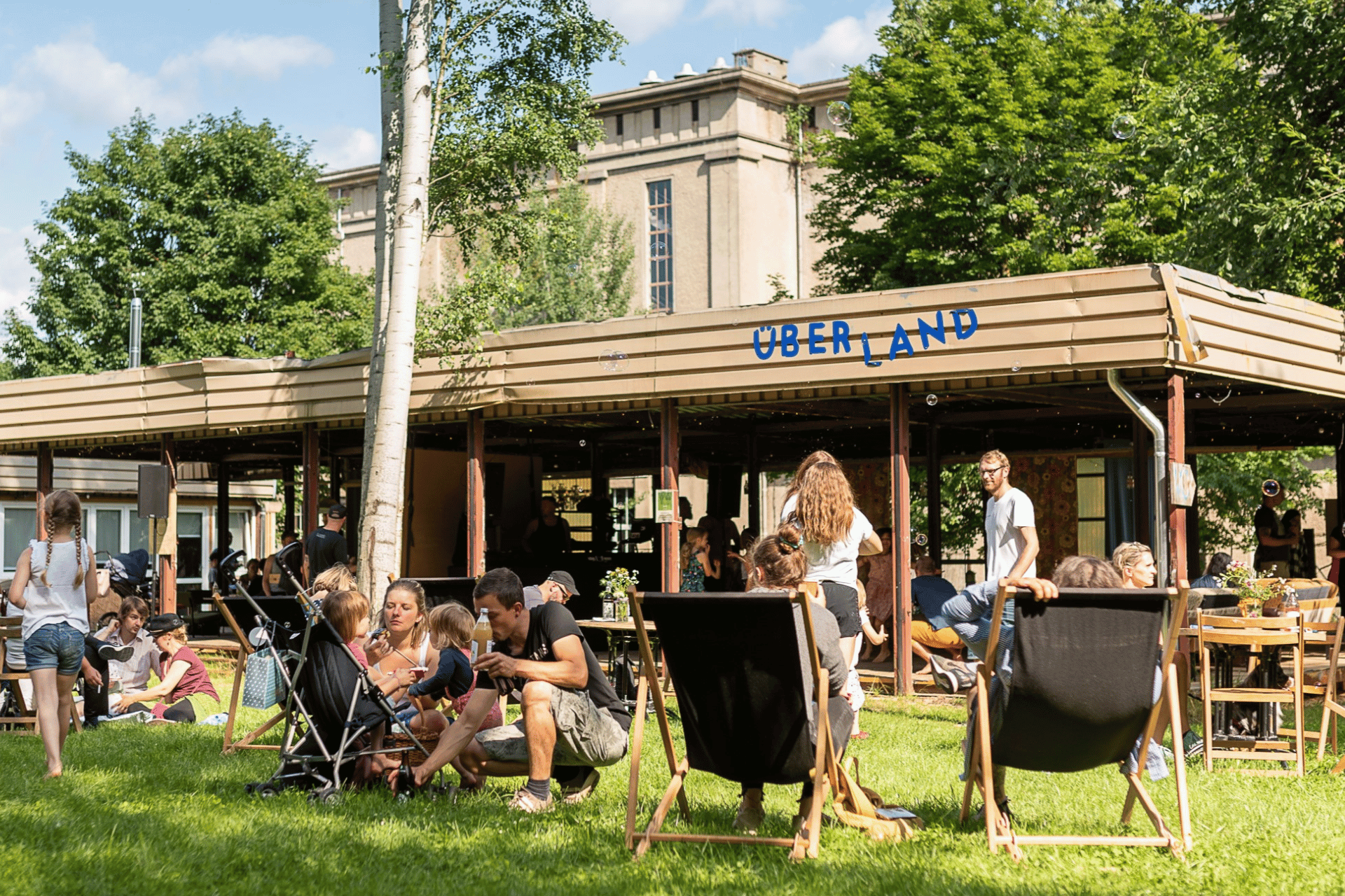 Ein sonniger Sommertag im Außenbereich einer Location namens „ÜBERLAND“. Im Vordergrund entspannen Menschen auf einer grünen Wiese; links sitzt eine Familie bei einem Picknick neben einem Kinderwagen, rechts ruhen Personen in Liegestühlen. Im Hintergrund befindet sich ein flaches, überdachtes Holzgebäude mit dem blauen Schriftzug „ÜBERLAND“ auf dem Dachsims, unter dem weitere Gäste im Schatten sitzen. Hinter dem Gelände ragen hohe grüne Bäume und die massive Fassade eines großen, grauen Industriegebäudes auf. Die Atmosphäre ist belebt und familiär, vereinzelt schweben Seifenblasen durch die Luft.