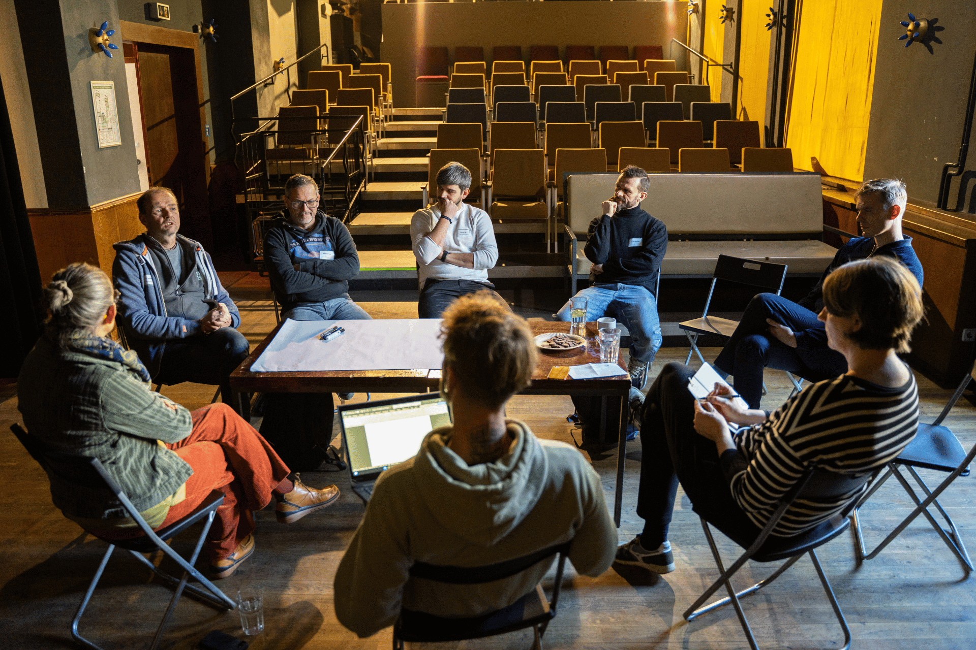 Eine Gruppe von etwa acht Personen sitzt in einem lockeren Kreis in einem abgedunkelten Veranstaltungsraum oder Theater. Sie sind in ein intensives Gespräch vertieft; einige Teilnehmer machen sich Notizen oder arbeiten an einem Laptop, während auf einem Tisch in der Mitte ein großes, leeres Blatt Papier und Stifte bereitliegen. Im Hintergrund sind aufsteigende Reihen von leeren Kinosesseln zu sehen, die durch warmes Licht sanft beleuchtet werden. Die Atmosphäre wirkt konzentriert und kooperativ, wie bei einem Workshop oder einer Planungsrunde.