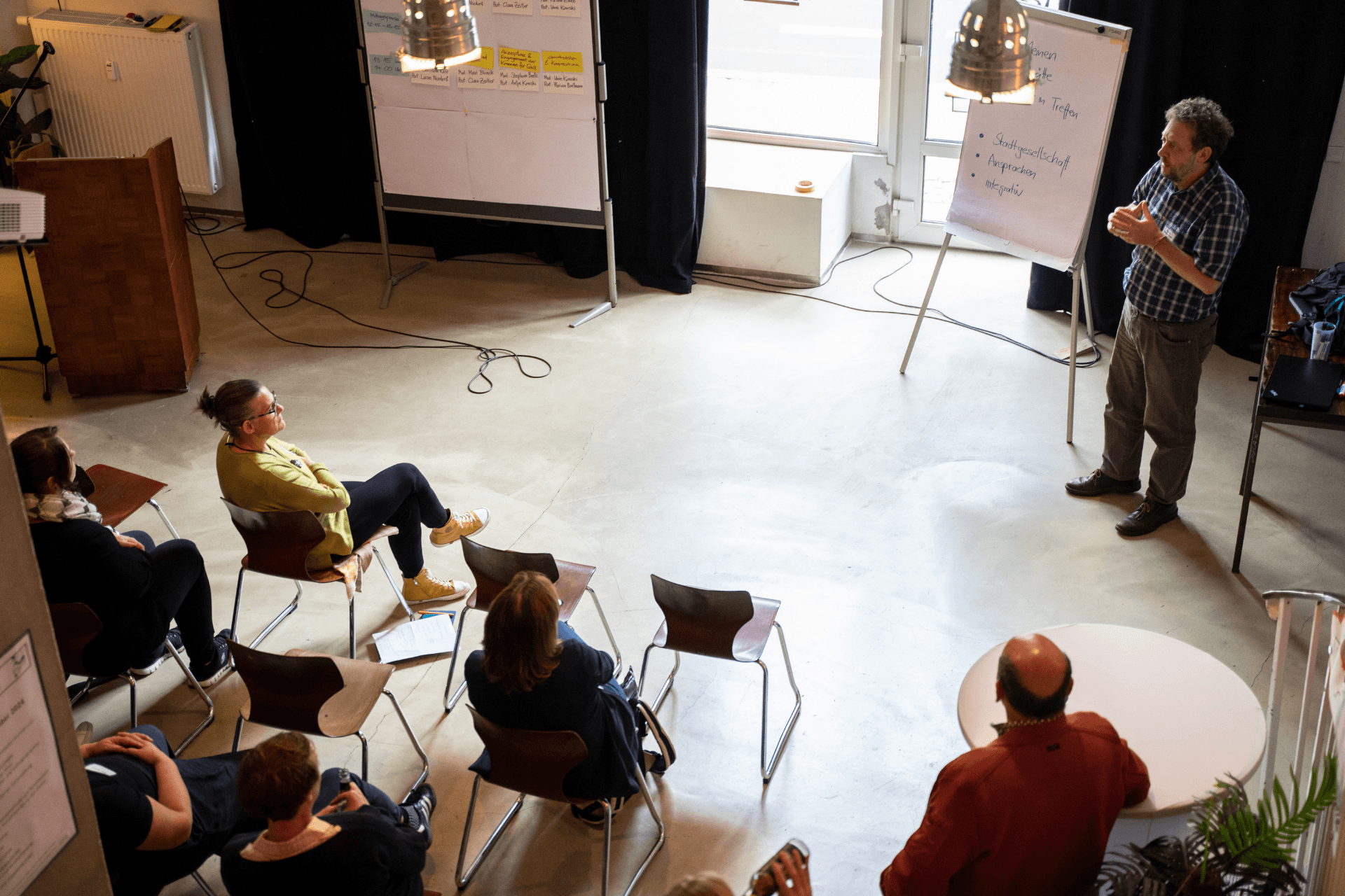 Eine Innenaufnahme aus erhöhter Perspektive, die eine Workshop-Situation zeigt. Ein Mann in einem karierten Hemd steht rechts vor einem Flipchart und erklärt der Gruppe seine Notizen. Vor ihm sitzen fünf Teilnehmerinnen und Teilnehmer halbkreisförmig auf einfachen Stühlen und hören aufmerksam zu. Im Hintergrund ist eine weitere große Moderationswand mit bunten Karteikarten zu sehen. Der Raum ist hell und wirkt durch das minimalistische Design wie ein modernes Atelier oder ein Seminarraum.