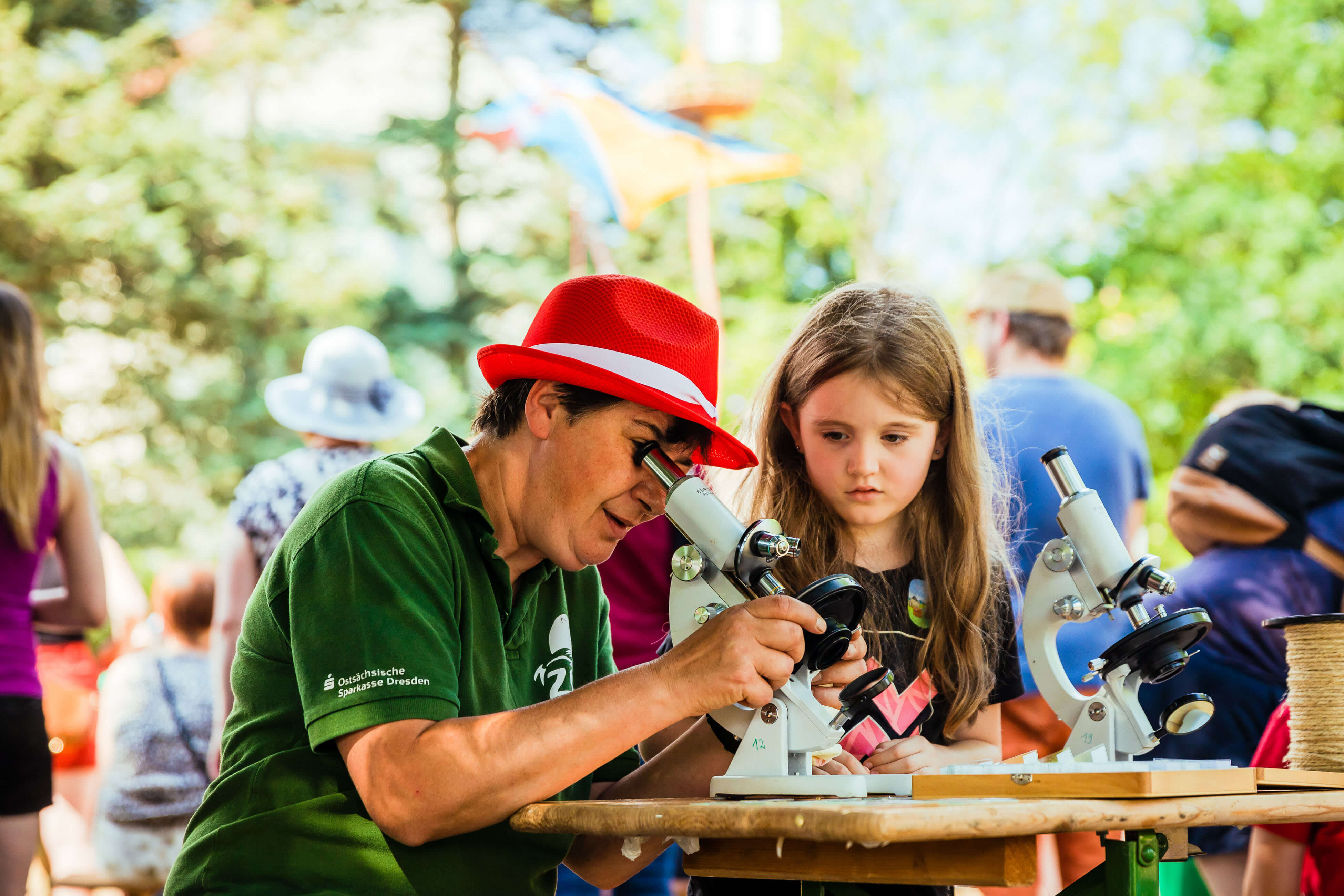  Gemini hat gesagt Person mit rotem Hut und Kind schauen gemeinsam durch ein Mikroskop auf einem Sommerfest im Freien.