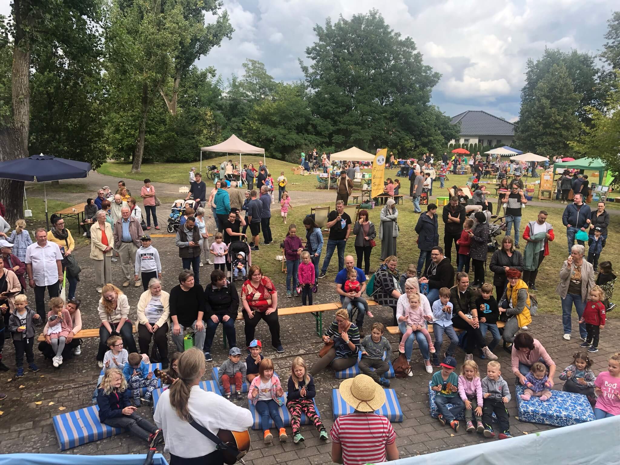 Zuschauer sitzen bei einem Open-Air-Kinderkonzert auf Bänken und Kissen im grünen Außenbereich.