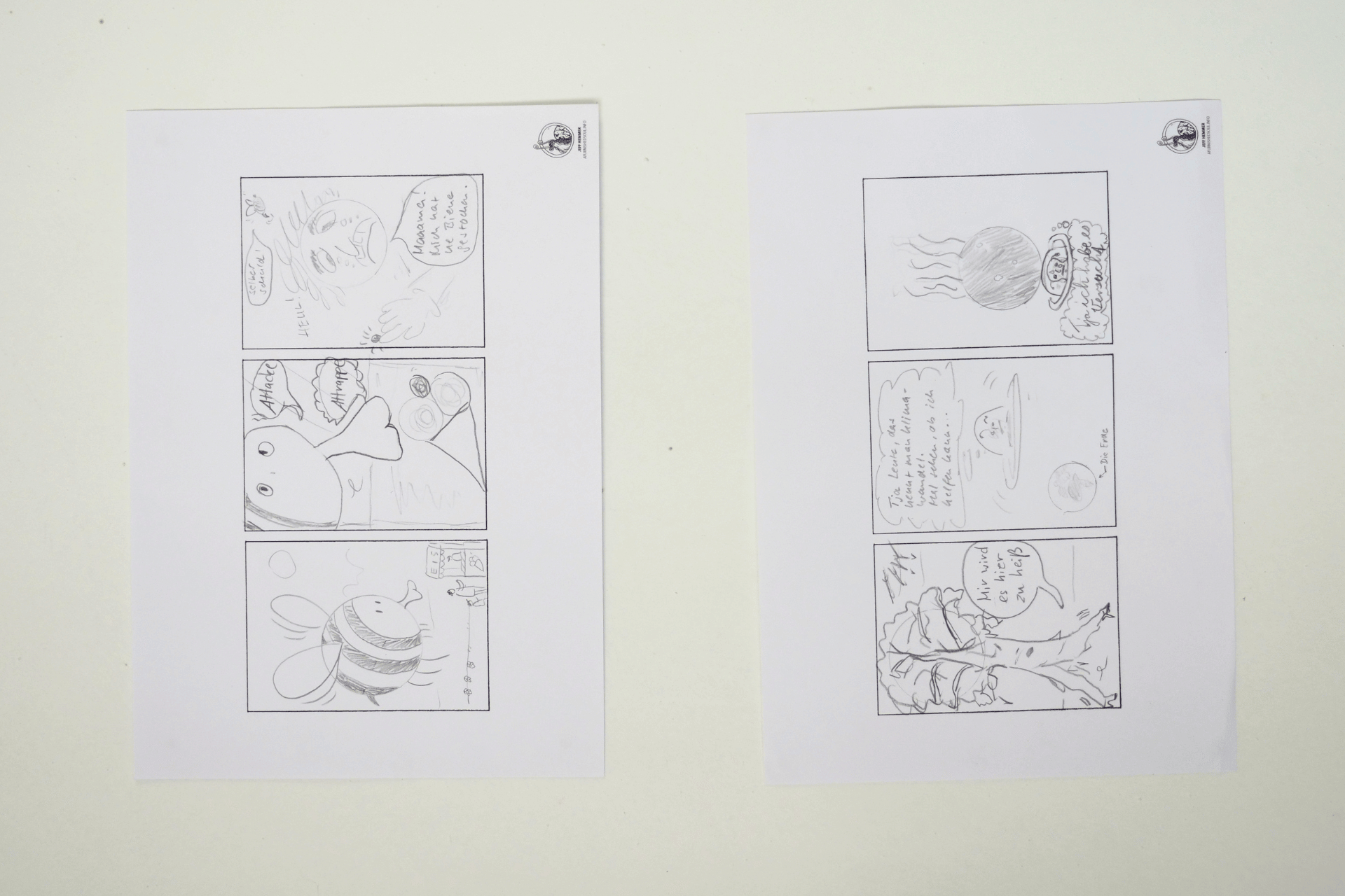 Zwei weiße Blätter mit handgezeichneten Comic-Seiten liegen nebeneinander auf einem hellen Tisch. Jedes Blatt zeigt drei schwarz-weiße Bleistift-Panels mit Figuren, Sprechblasen und skizzenhaften Szenen.