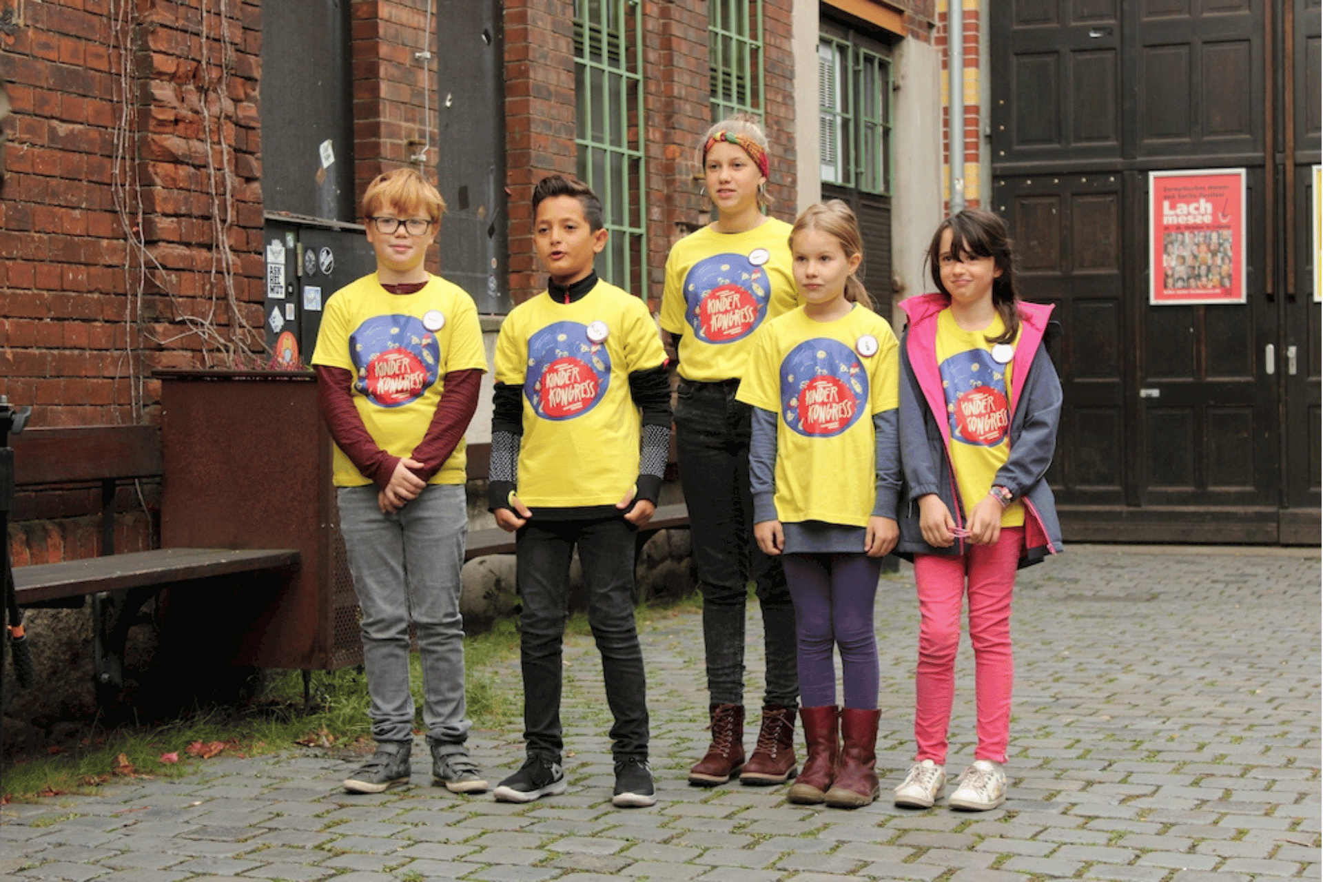 Eine Gruppe von fünf Kindern steht in einer Reihe auf einem gepflasterten Platz vor einem Backsteingebäude. Alle Kinder tragen gelbe T-Shirts mit dem Aufdruck „Kinderkongress“. Sie schauen in Richtung der Kamera. Im Hintergrund ist eine große dunkle Holztür sowie ein Schild mit der Aufschrift „Lachmesse“ zu sehen.