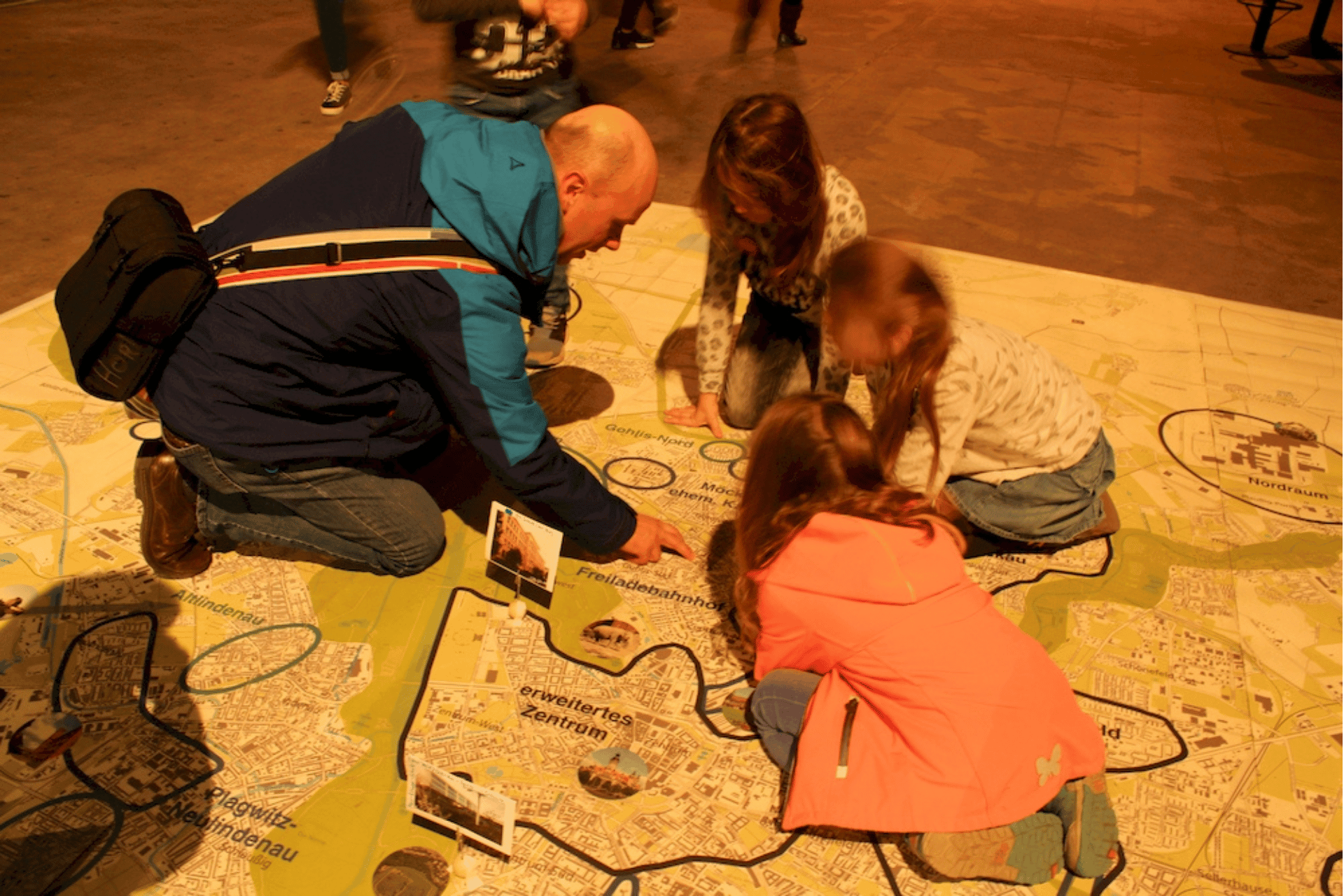 Ein Erwachsener kniet gemeinsam mit drei Kindern auf dem Boden, um eine große, auf dem Boden ausgebreitete Landkarte zu betrachten. Die Karte zeigt verschiedene Stadtgebiete und ist mit kleinen Modellen oder Markierungen versehen, die bestimmte Orte kennzeichnen. Die Gruppe scheint sich intensiv über die Karte auszutauschen und gemeinsam einen Ort zu suchen oder zu planen.