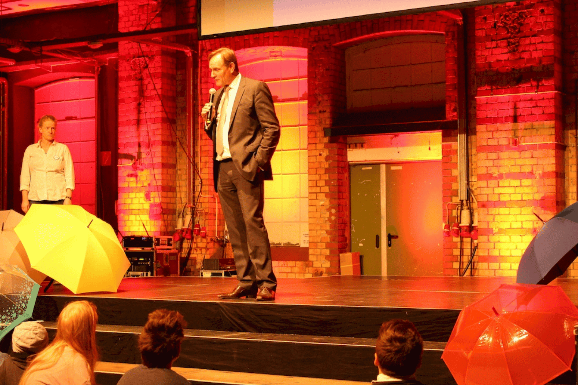 Ein Mann in einem dunklen Anzug steht auf einer Bühne und spricht in ein Mikrofon. Die Bühne ist durch eine auffällige rote Beleuchtung in Szene gesetzt, im Hintergrund ist eine Wand aus Backsteinen zu sehen. Im Vordergrund sind die Köpfe von Zuschauern erkennbar, und auf der Bühne stehen oder liegen mehrere bunte Regenschirme.