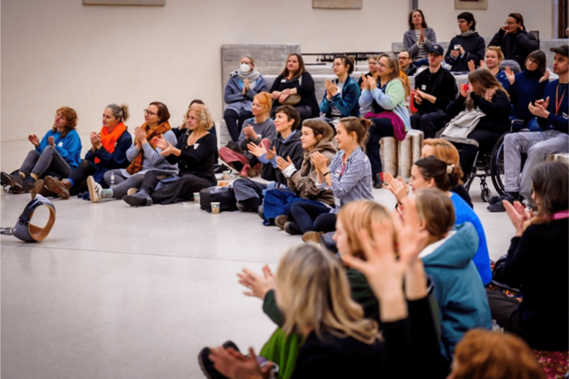 Eine große Gruppe von Menschen sitzt in einem weiten, hellen Raum auf dem Boden und auf Podesten. Fast alle klatschen Beifall und blicken aufmerksam in Richtung eines nicht im Bild sichtbaren Geschehens. Die Stimmung wirkt begeistert und gemeinschaftlich. Im Vordergrund sind einige Personen nur unscharf von hinten zu sehen, während die Reihen dahinter klar erkennbar sind. Der Raum ist schlicht gestaltet und bietet viel freie Fläche.