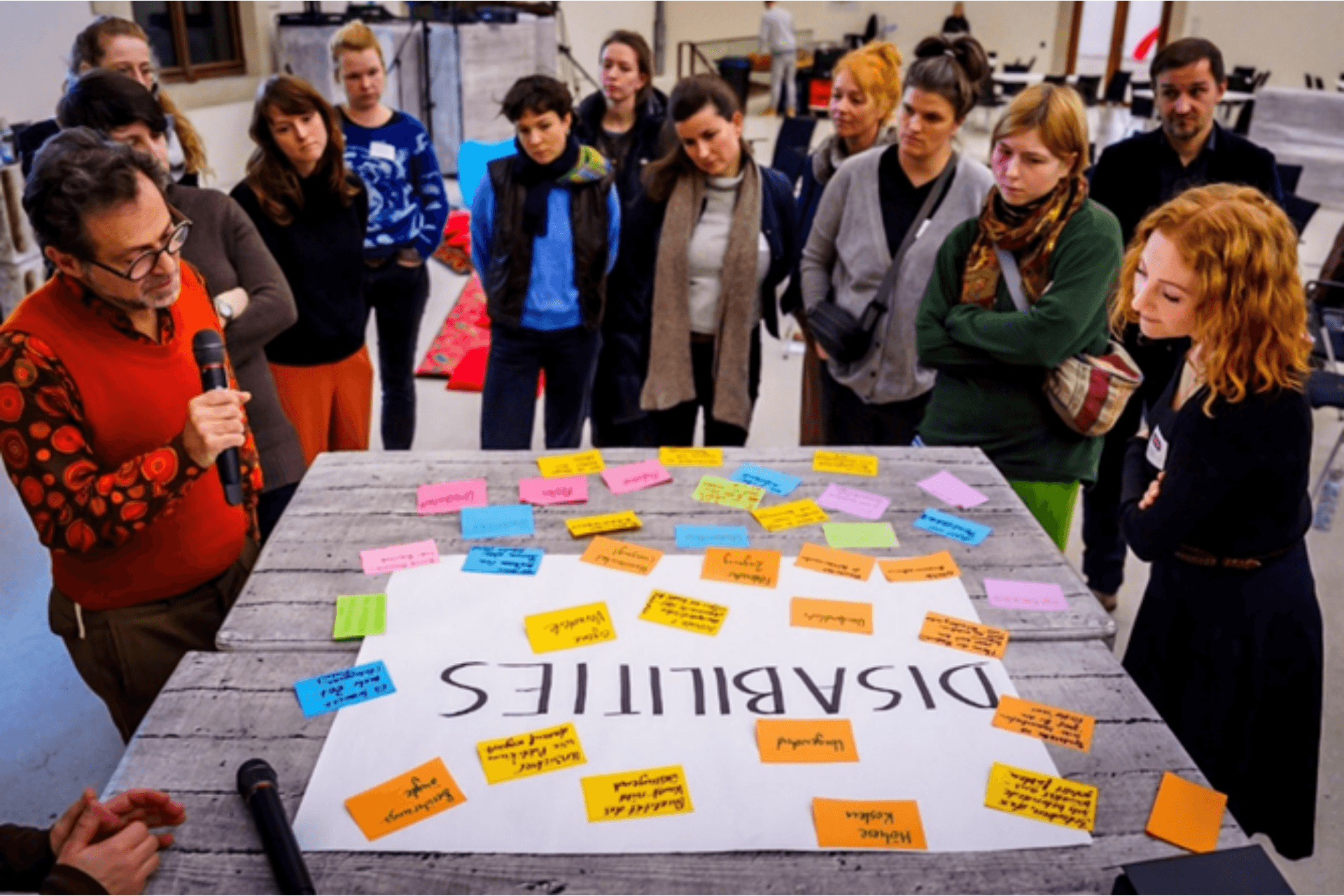 Eine Gruppe von etwa 15 Personen steht in einem hellen Raum um einen großen Arbeitstisch herum. Auf dem Tisch liegt ein weißes Plakat mit der zentralen Aufschrift „DISABILITIES“, das mit zahlreichen bunten Klebezetteln (Post-its) beklebt ist. Ein Mann auf der linken Seite spricht in ein Mikrofon und erläutert die gesammelten Punkte, während die anderen Teilnehmer aufmerksam auf das Plakat schauen oder dem Sprecher zuhören. Die Szene zeigt eine aktive Workshop-Phase oder Präsentation zum Thema Inklusion und Barrierefreiheit.