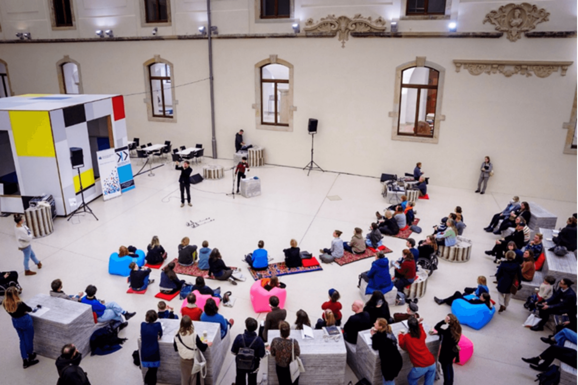 Eine Weitwinkelaufnahme aus erhöhter Perspektive auf eine Veranstaltung in einem großen, hellen Lichthof mit historischer Architektur. Im Zentrum steht eine Person vor einem Mikrofon und spricht zu einem Publikum von etwa 50 bis 60 Personen. Die Zuschauer sitzen locker verteilt auf dem Boden, auf bunten Sitzsäcken oder auf grauen, quaderförmigen Sitzelementen. Links im Bild steht eine große, kubische Installation im Stil von Piet Mondrian mit gelben, roten und blauen Flächen. Die hohen weißen Wände sind durch Rundbogenfenster und klassische Steinornamente gegliedert. Die Szene wirkt offen, modern und kommunikativ.