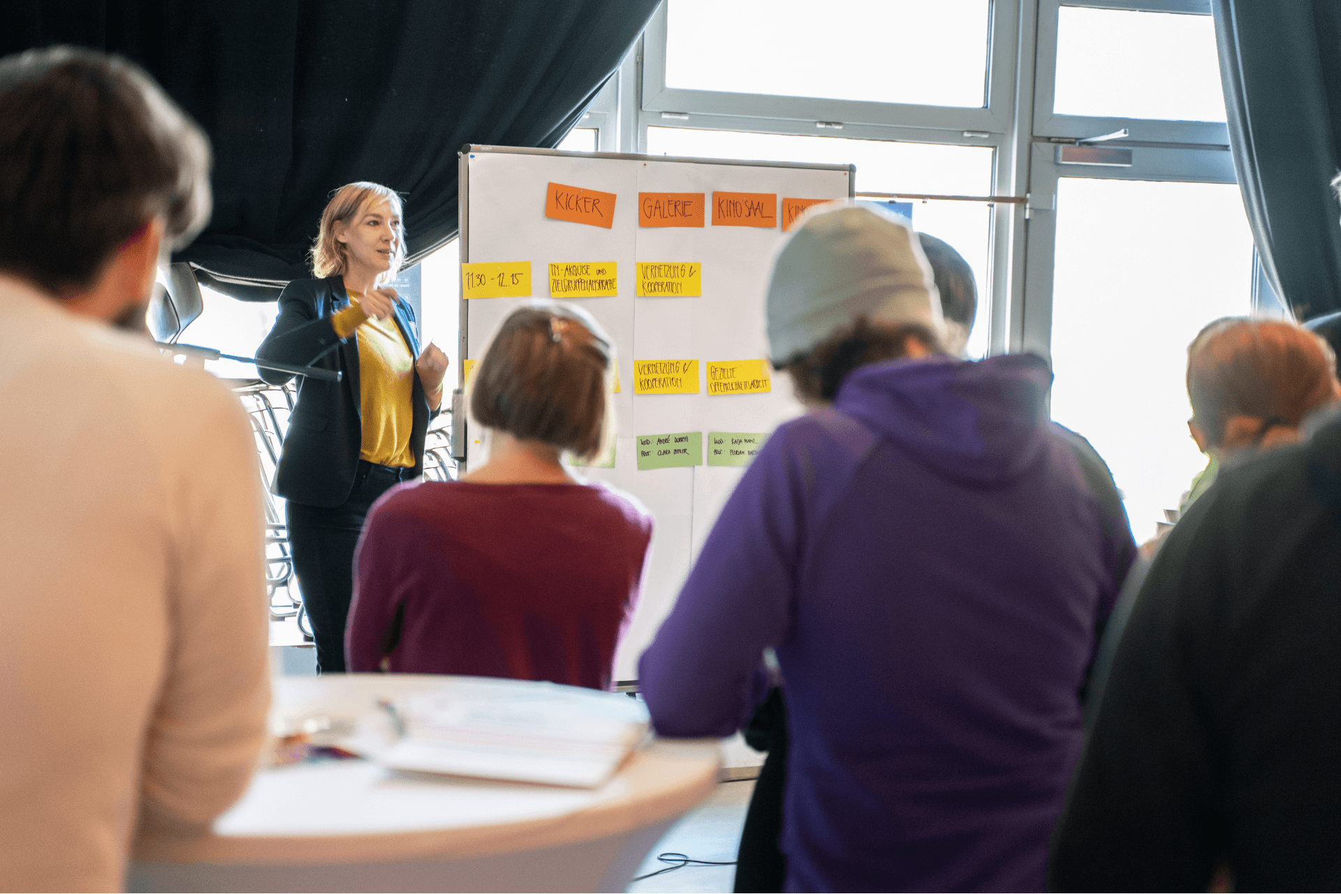 Eine Workshop-Situation in einem hellen Raum. Eine Frau mit blonden Haaren, die eine gelbe Bluse und einen dunklen Blazer trägt, steht vor einem Flipchart und spricht gestikulierend zu einer Gruppe. Auf dem Flipchart sind verschiedene bunte Moderationskarten angebracht, unter anderem mit den Überschriften „KICKER“, „GALERIE“ und „KINOSAAL“. Im Vordergrund sind die Rückenansichten mehrerer Teilnehmer zu sehen, die aufmerksam der Präsentation folgen. Große Fenster im Hintergrund lassen viel Tageslicht in den Raum.