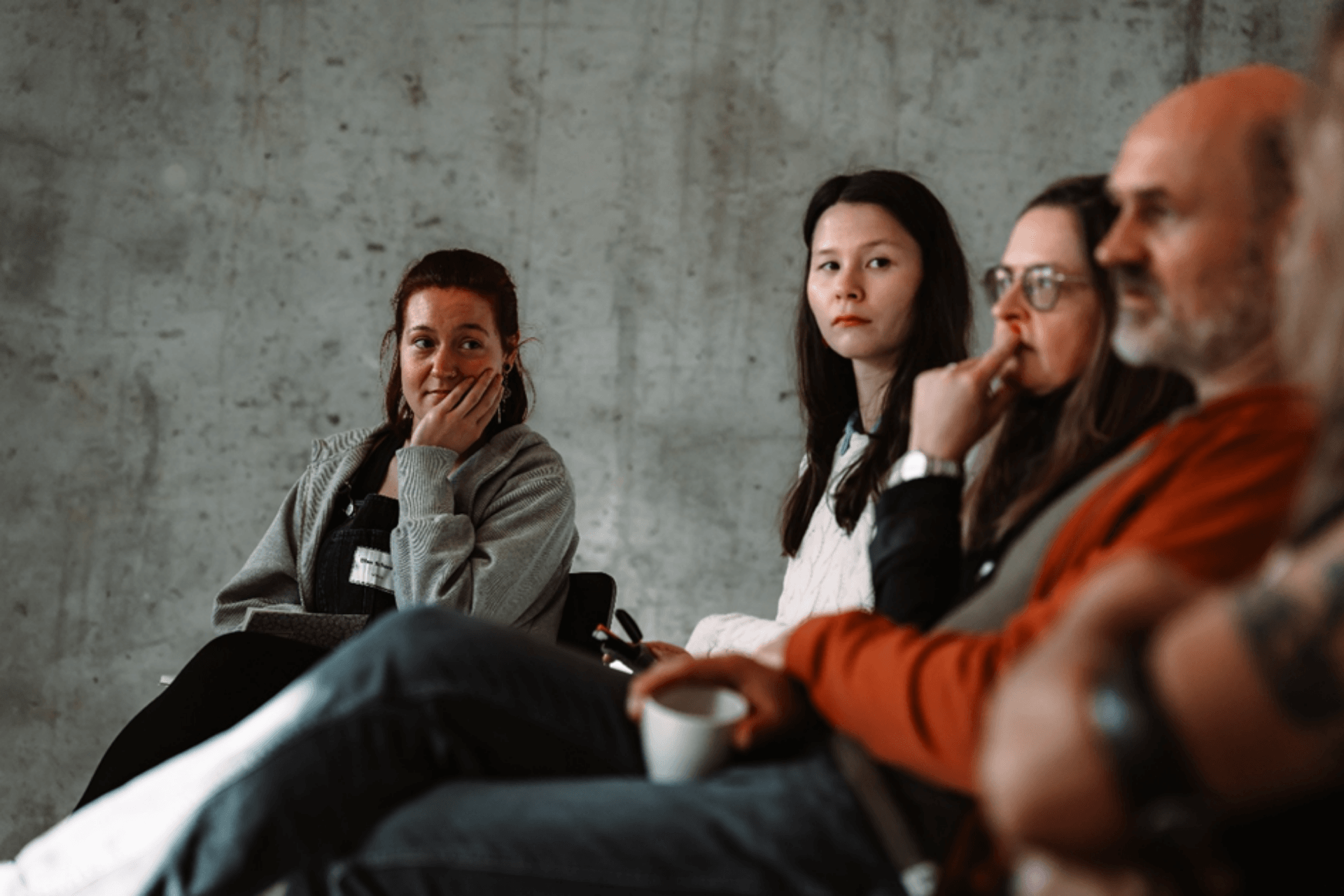 Eine Gruppe von vier Personen sitzt nebeneinander vor einer grauen Sichtbetonwand und hört aufmerksam zu. Im Fokus steht eine Frau auf der linken Seite mit rötlichem Haar und einer grauen Strickjacke, die ihren Kopf nachdenklich auf ihre Hand stützt. Rechts daneben sitzen zwei weitere Frauen und ein Mann im Profil, die konzentriert in dieselbe Richtung blicken. Die Szene wirkt ruhig und reflektiert, wie während eines Vortrags oder einer Diskussionsrunde.