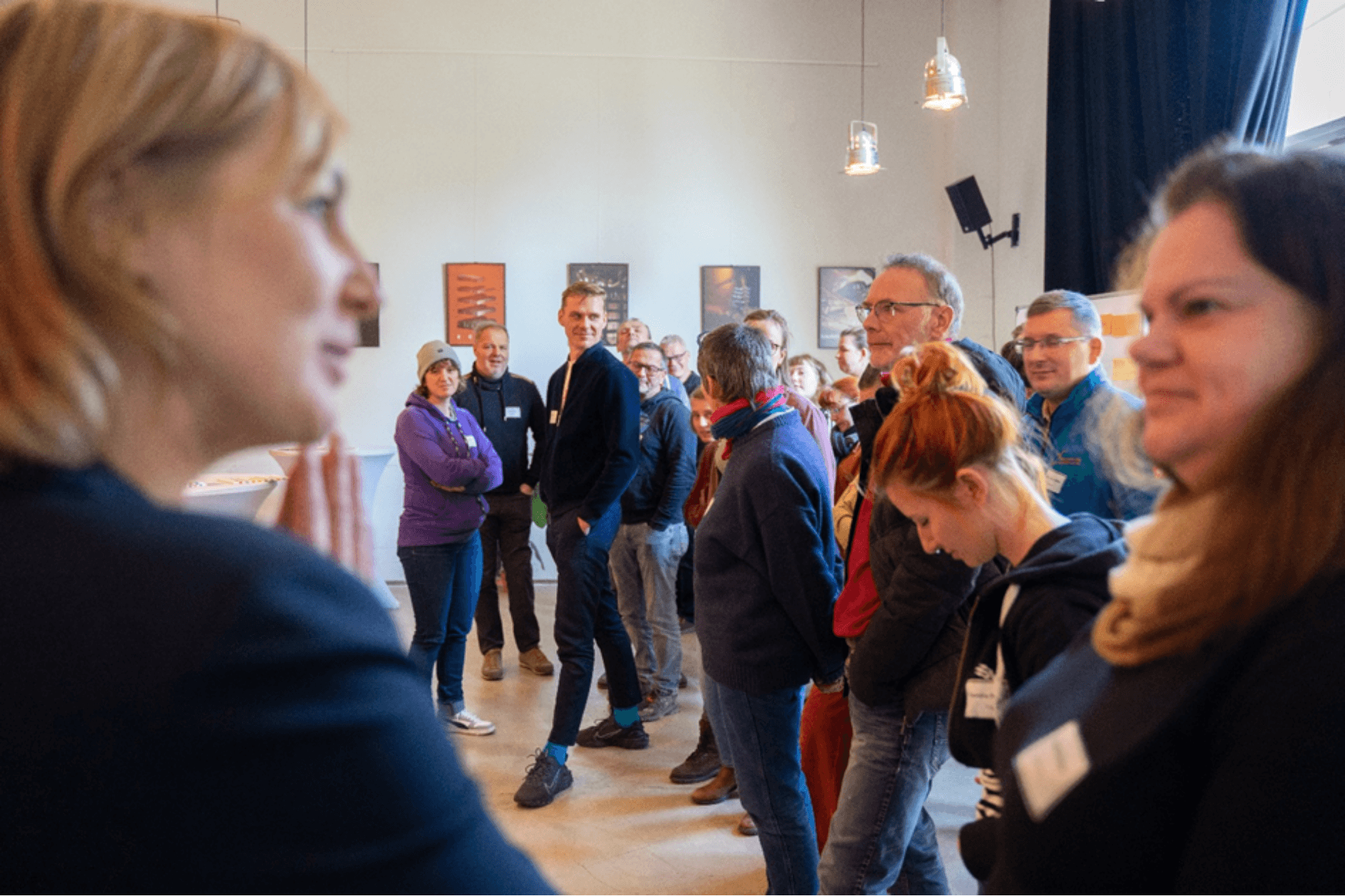 Eine lebendige Szene während einer Veranstaltung in einem hellen Raum. Im Vordergrund sind zwei Frauen im Profil zu sehen, die den Bildrand einrahmen und den Blick auf eine Gruppe von etwa zehn Personen im Mittelgrund lenken. Diese Teilnehmer stehen locker zusammen und blicken interessiert in Richtung der Sprecherin links im Vordergrund. An der weißen Wand im Hintergrund hängen mehrere gerahmte Bilder in einer Reihe, und moderne Industrie-Hängelampen beleuchten den Raum. Die Atmosphäre wirkt aufgeschlossen und erwartungsvoll.