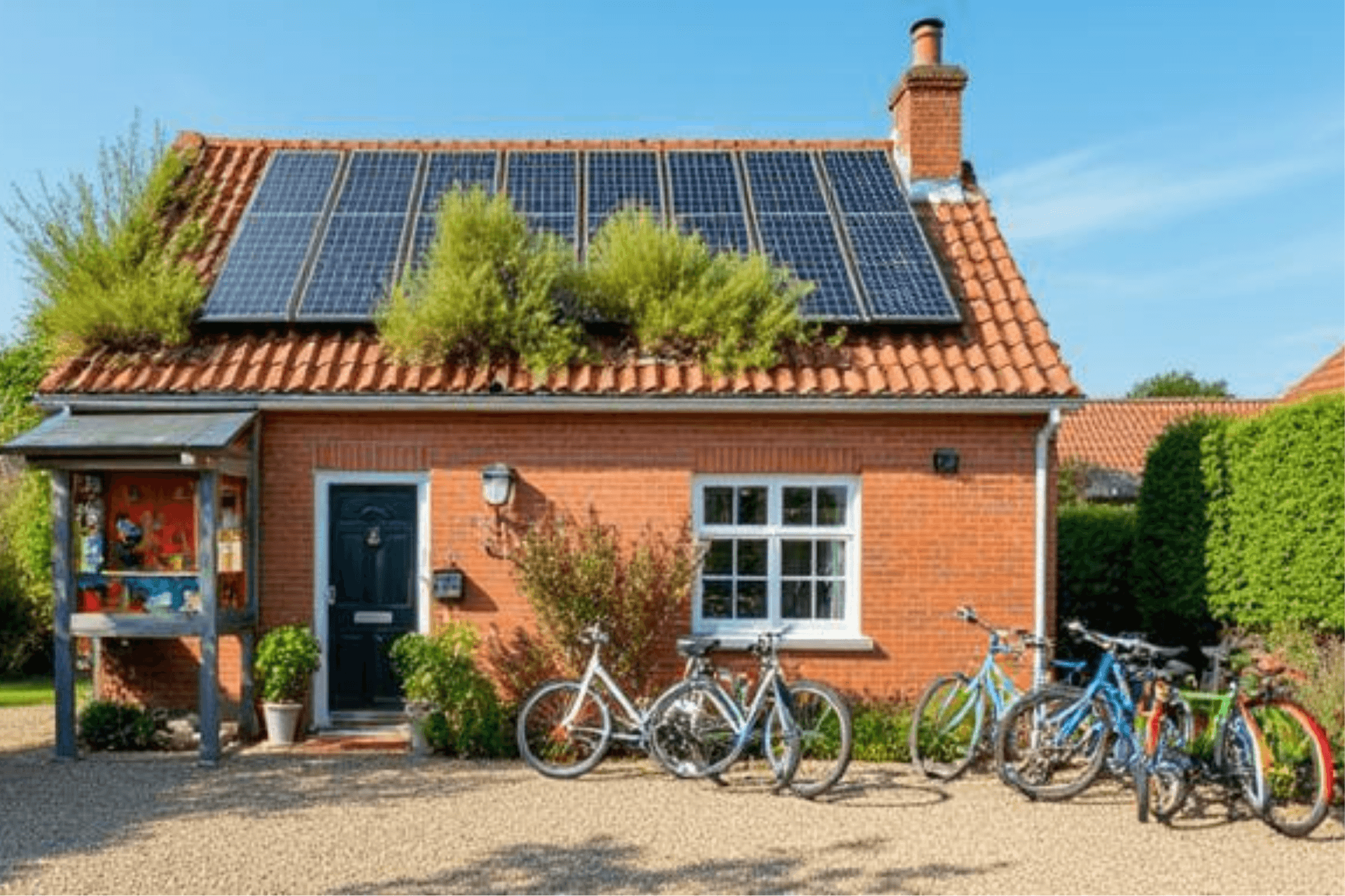 Ein kleines, einstöckiges Backsteinhaus unter blauem Himmel. Auf dem roten Ziegeldach ist eine große Photovoltaikanlage installiert, zwischen deren Paneelen und am Dachrand üppige grüne Pflanzen wachsen. Vor dem Haus stehen mehrere Fahrräder in verschiedenen Farben an die Wand gelehnt. Links neben der dunkelblauen Eingangstür befindet sich ein kleiner, überdachter Schaukasten auf Holzpfosten, der bunt gestaltet ist. Die Szene wirkt idyllisch, ökologisch orientiert und einladend.