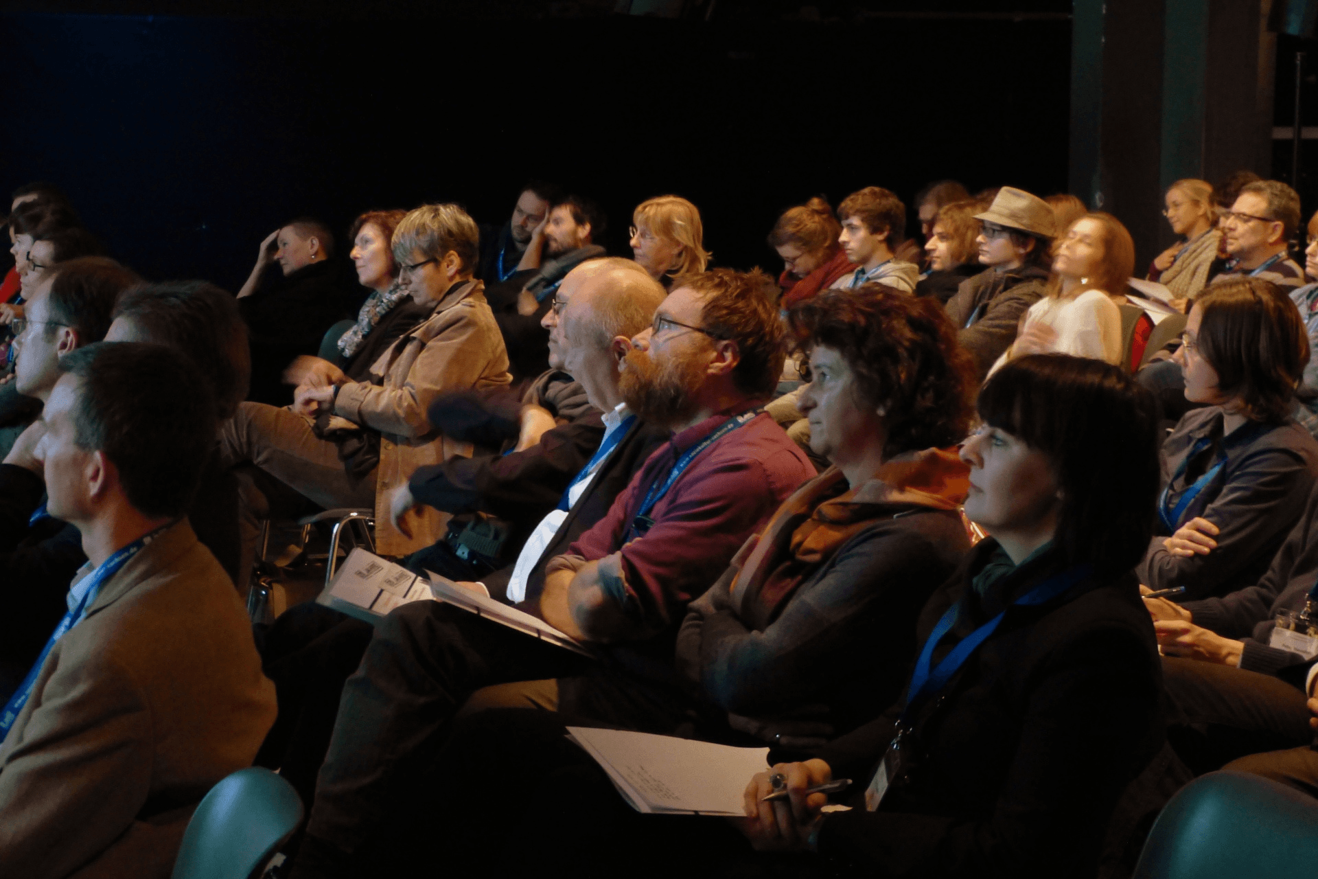 Eine Seitenansicht eines Publikums in einem abgedunkelten Veranstaltungsraum. Zahlreiche Personen sitzen dicht nebeneinander in Stuhlreihen und blicken konzentriert in Richtung einer Bühne. Viele Teilnehmer tragen blaue Schlüsselbänder (Lanyards) um den Hals und halten Infomaterialien oder Notizblöcke auf dem Schoß. Die Beleuchtung ist gedämpft und fokussiert auf die Gesichter der Zuhörer, was eine aufmerksame und ruhige Tagungsatmosphäre vermittelt.
