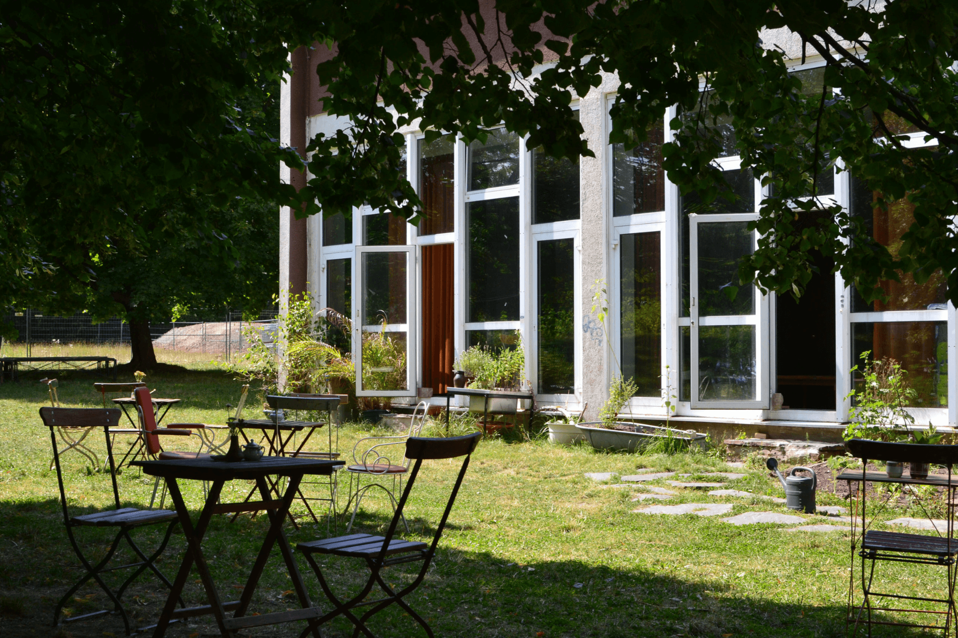 Ein sonniger Außenbereich eines Gebäudes mit einer großen Glasfassade, die aus hohen, weiß gerahmten Fenstern und Türen besteht. Davor erstreckt sich eine grüne Wiese, auf der mehrere dunkle Klapptische und Metallstühle im Schatten großer Bäume verteilt stehen. Die Szene wirkt wie ein einladendes Gartencafé oder ein Pausenbereich im Freien. Vereinzelt stehen Pflanzenkübel und eine Gießkanne an der Hauswand, während im Hintergrund eine sonnenbeschienene Rasenfläche und weitere Bäume zu sehen sind.