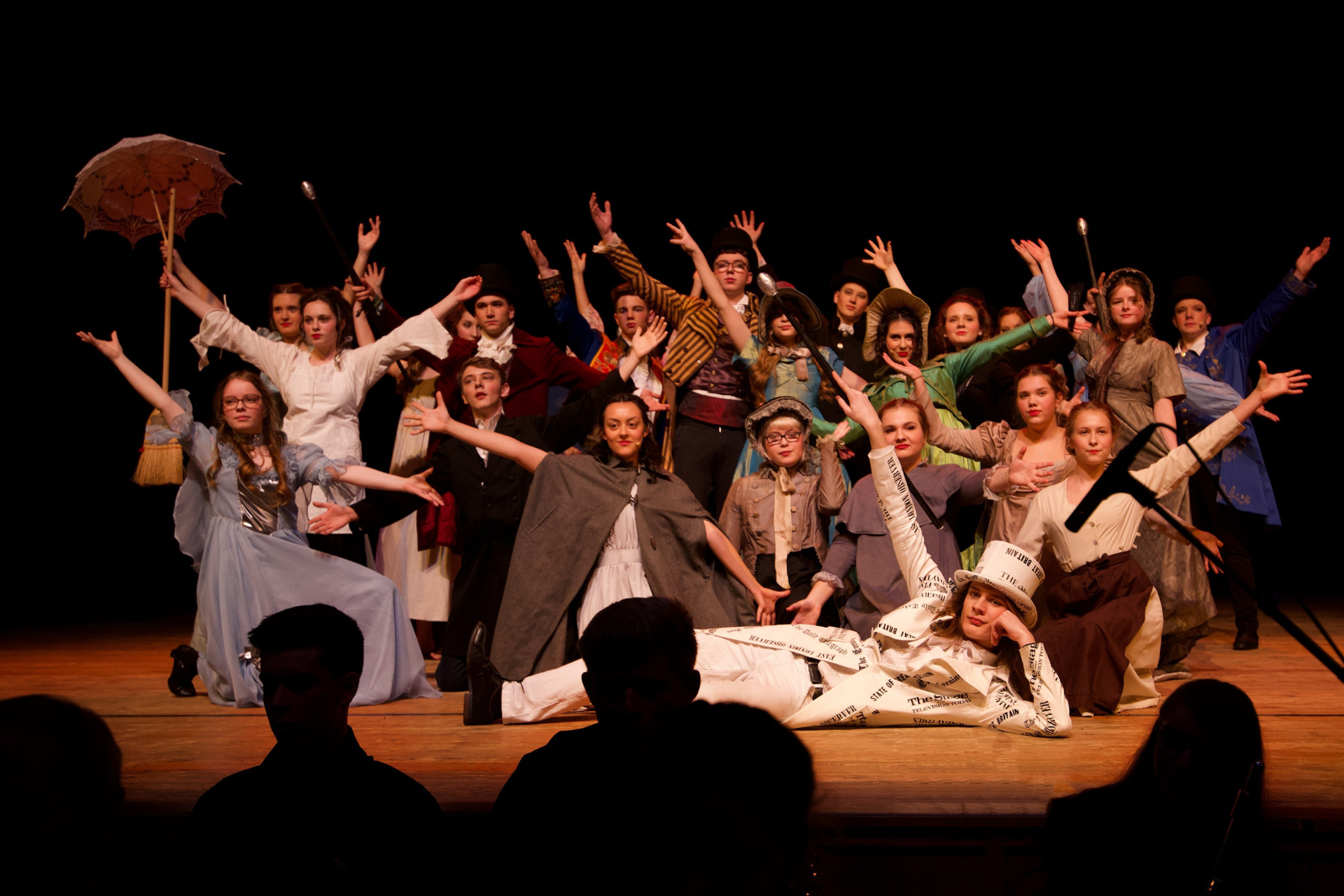  Eine lebendige Theateraufführung mit einer großen Gruppe von Schauspielern in historischer Kostümierung. Die Darsteller stehen in verschiedenen Posen auf der Bühne, einige heben die Arme in die Luft, während andere auf dem Boden liegen oder lächeln. Die Beleuchtung hebt die farbenfrohen Kostüme hervor, und der Zuschauerraum ist teilweise sichtbar, mit Silhouetten von Menschen im Vordergrund.