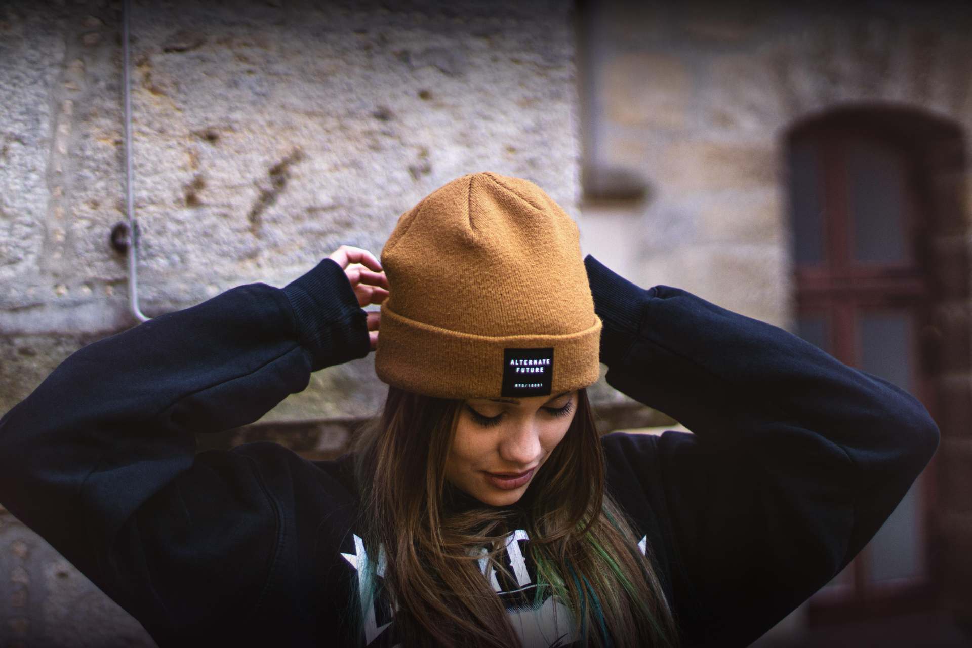 Eine junge Frau mit langem, brünettem Haar trägt eine camel-farbene Mütze und ein schwarzes Sweatshirt. Sie schaut nach unten und passt ihre Mütze an, während sie vor einer unscharfen, steinernen Wand steht.