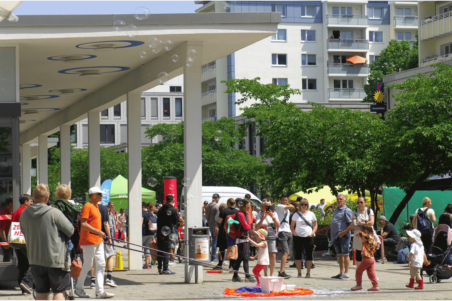 Eine belebte Straßenszene auf einem öffentlichen Platz vor einem modernen Wohngebäude. Im Vordergrund erzeugt ein Mann mit großen Stäben riesige Seifenblasen, die durch die Luft schweben und die Aufmerksamkeit von Passanten und Kindern auf sich ziehen. Zahlreiche Menschen spazieren über den Platz oder bleiben stehen, um zuzuschauen. Im Hintergrund sind bunte Marktzelte, grüne Bäume und die Fassade eines mehrstöckigen Wohnhauses mit Balkonen zu sehen. Die Szene vermittelt eine sommerliche, entspannte Stadtteilfest-Atmosphäre.