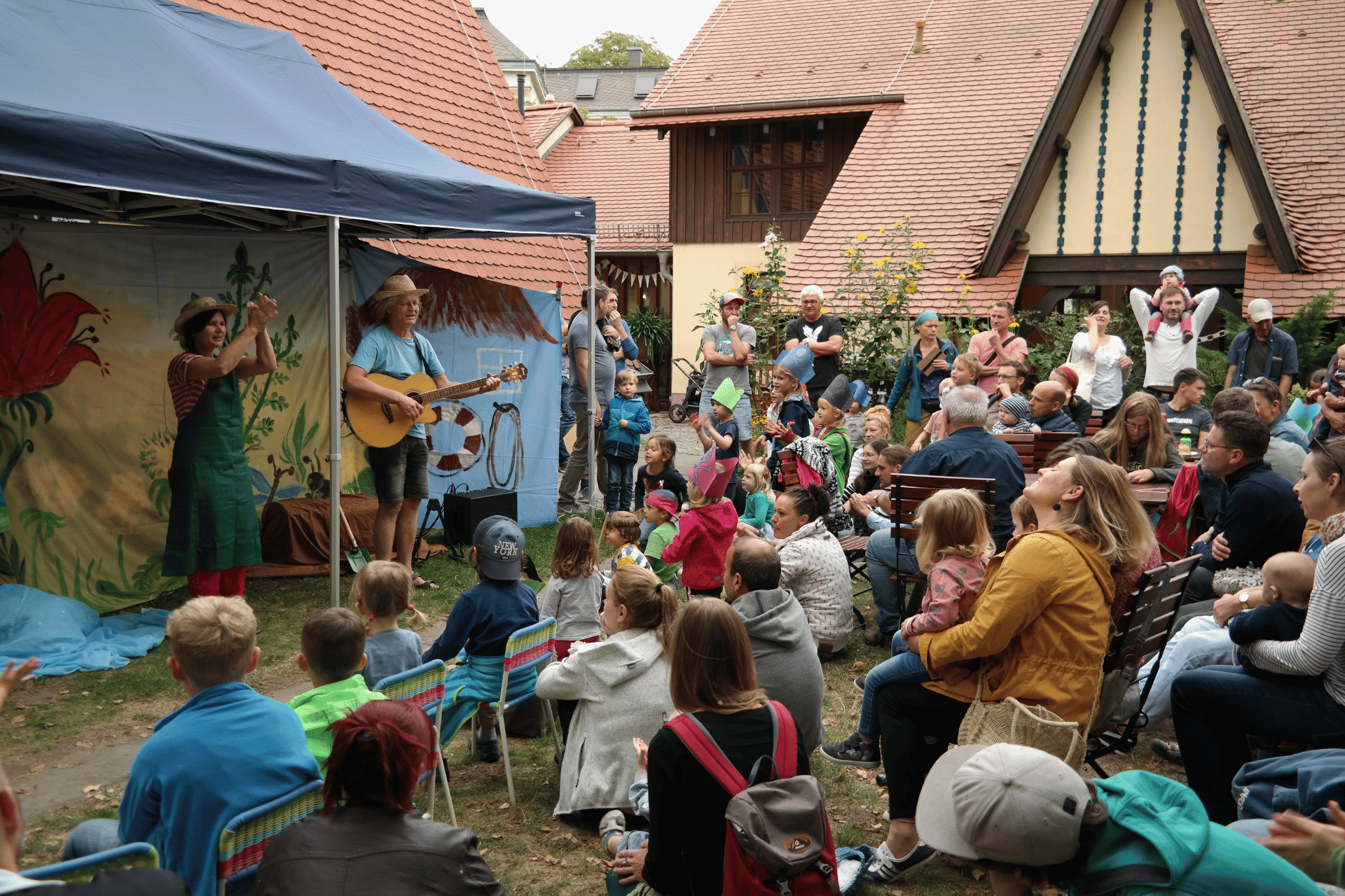 Eine sommerliche Open-Air-Veranstaltung in einem gemütlichen Innenhof mit Fachwerkhäusern und roten Ziegeldächern. Unter einem blauen Pavillon treten zwei Personen vor einer bunt bemalten Kulisse auf: Eine Frau in einer grünen Schürze klatscht im Rhythmus, während ein Mann daneben Gitarre spielt und singt. Ein großes Publikum, bestehend aus vielen Kindern und Erwachsenen, sitzt aufmerksam davor auf der Wiese oder auf Bierbänken. Einige Kinder tragen bunte, selbstgebastelte Papierkronen. Die Stimmung ist fröhlich und gemeinschaftlich.