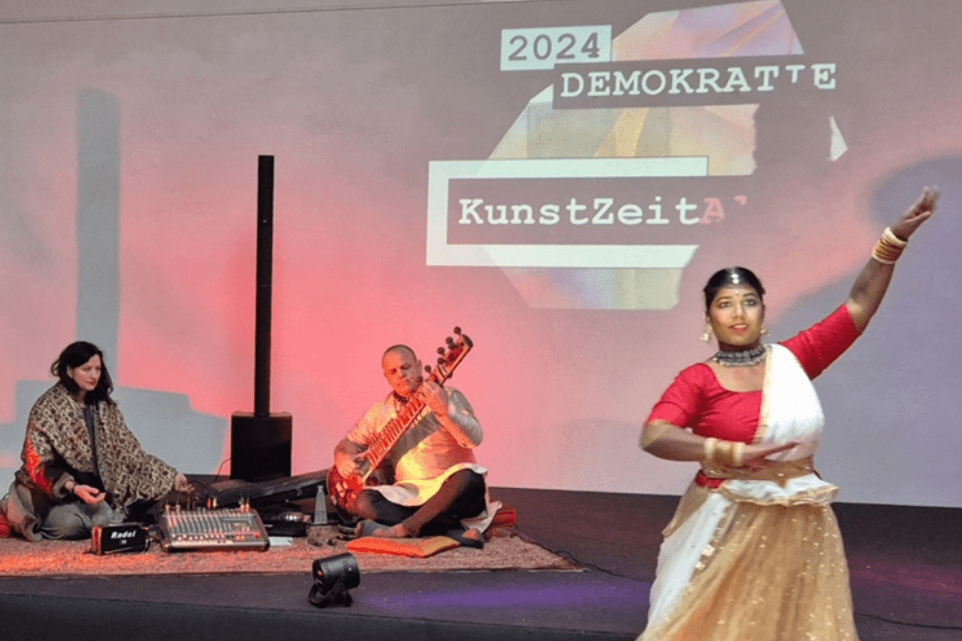 Eine Bühnenperformance während der Veranstaltung „KunstZeitAll 2024 DEMOKRATIE“. Im Vordergrund tanzt eine Frau in einem traditionellen indischen Gewand (Sari) in Rot, Weiß und Gold mit ausdrucksstarken Handbewegungen. Im Hintergrund sitzen zwei Musiker auf Teppichen: Ein Mann spielt eine Sitar und eine Frau bedient ein Mischpult sowie weitere elektronische Geräte. Die Bühne wird von rötlichem Scheinwerferlicht beleuchtet, und im Hintergrund ist eine Projektion mit dem Veranstaltungstitel zu sehen. Die Szene vermittelt eine Verbindung von traditioneller Kunst und moderner Performance.