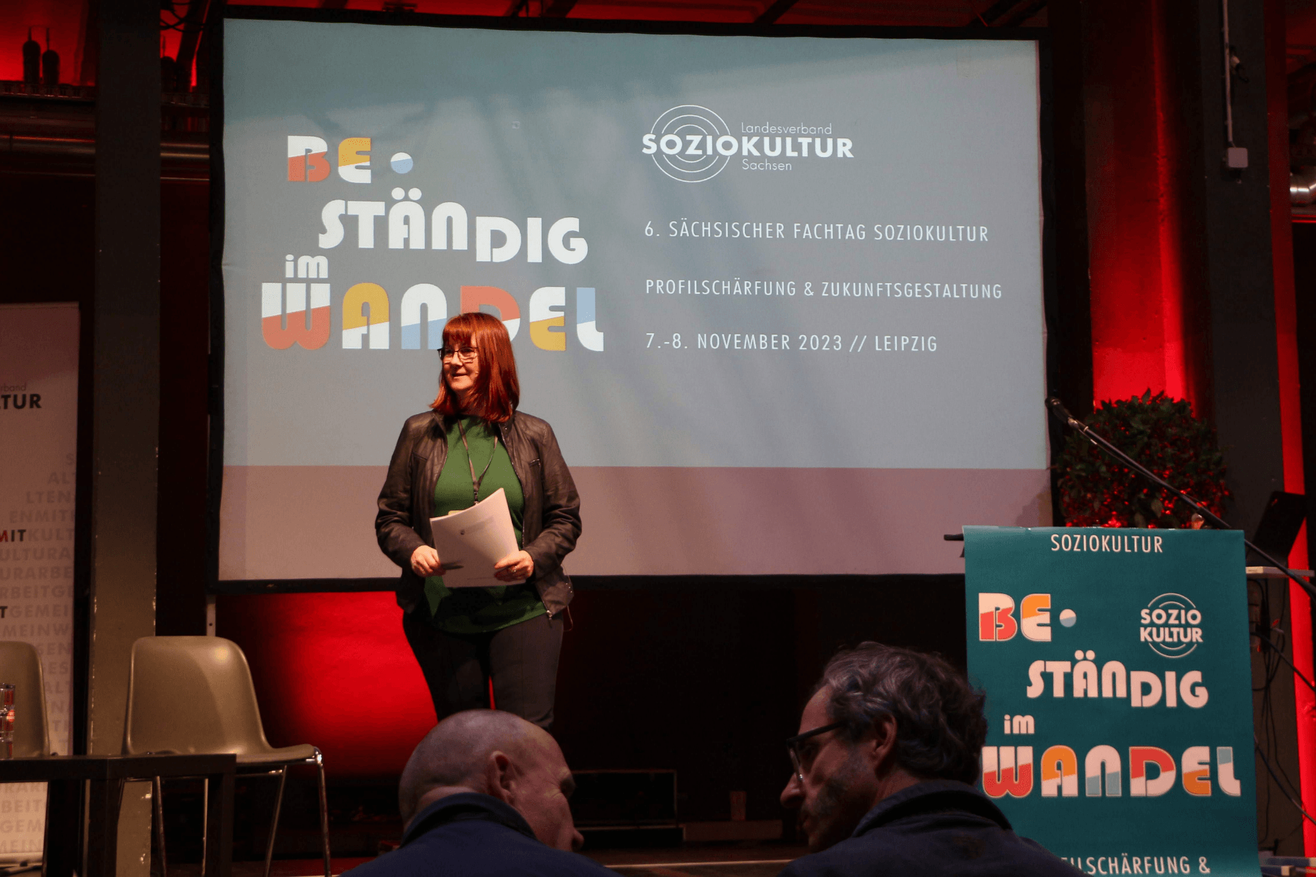 Eine Sprecherin mit roten Haaren und Brille steht auf einer Bühne vor einer großen Leinwand. Sie trägt eine dunkle Lederjacke über einem grünen Oberteil und hält Unterlagen in den Händen. Auf der Leinwand ist das Logo des „Landesverband Soziokultur Sachsen“ sowie der Titel der Veranstaltung zu sehen: „BE-STÄNDIG im WANDEL – 6. Sächsischer Fachtag Soziokultur“. Darunter stehen die Themen „Profilschärfung & Zukunftsgestaltung“ sowie das Datum „7.–8. November 2023 // Leipzig“. Im Vordergrund ist ein Rednerpult mit demselben Veranstaltungsdesign zu erkennen, und im Anschnitt sieht man die Köpfe zweier Zuhörer. Die Bühne ist stimmungsvoll mit rotem Licht hinterleuchtet.