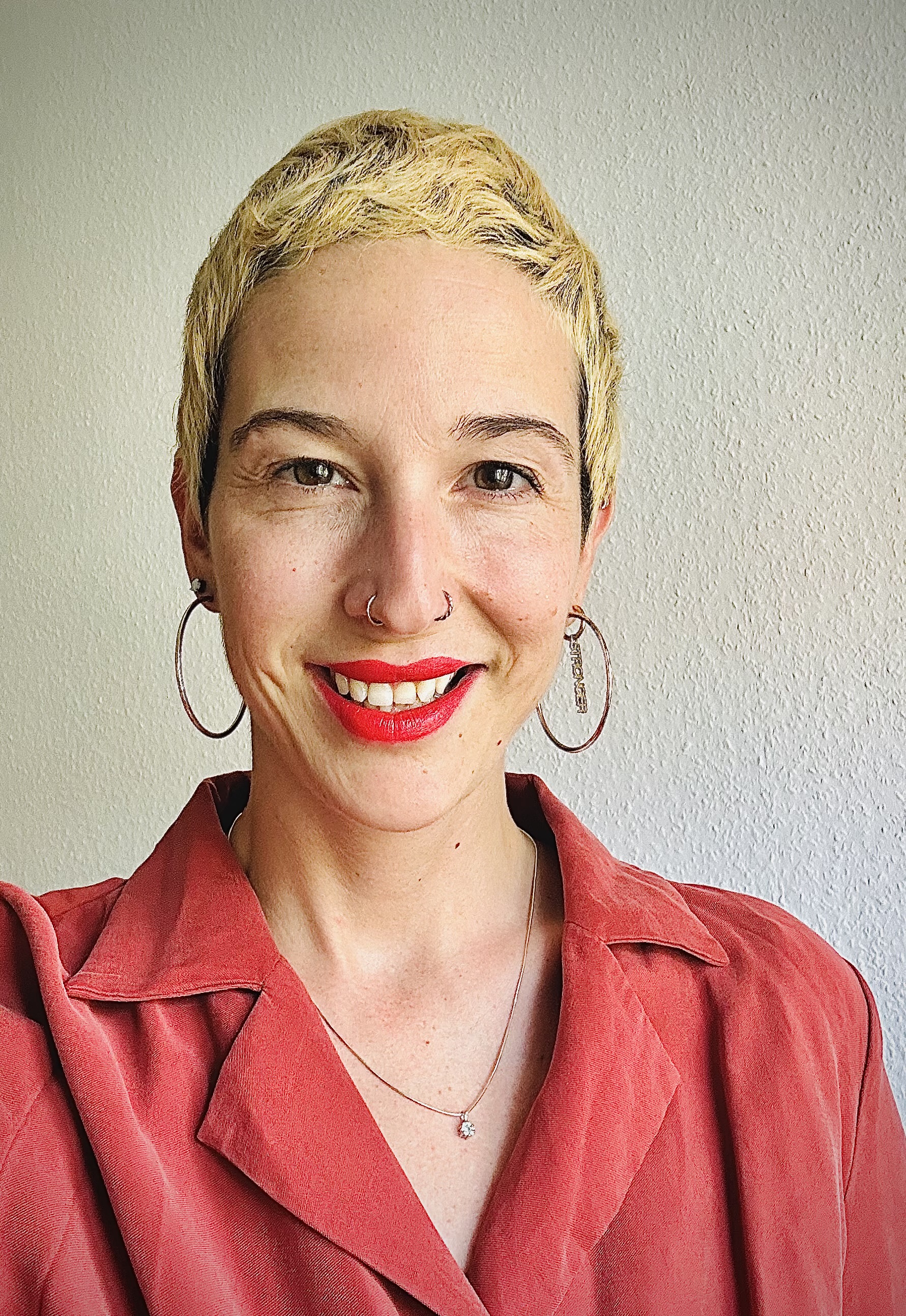 Ein Porträt einer lächelnden Frau mit kurzen blonden Haaren, rotem Lippenstift und großen Ohrringen.
