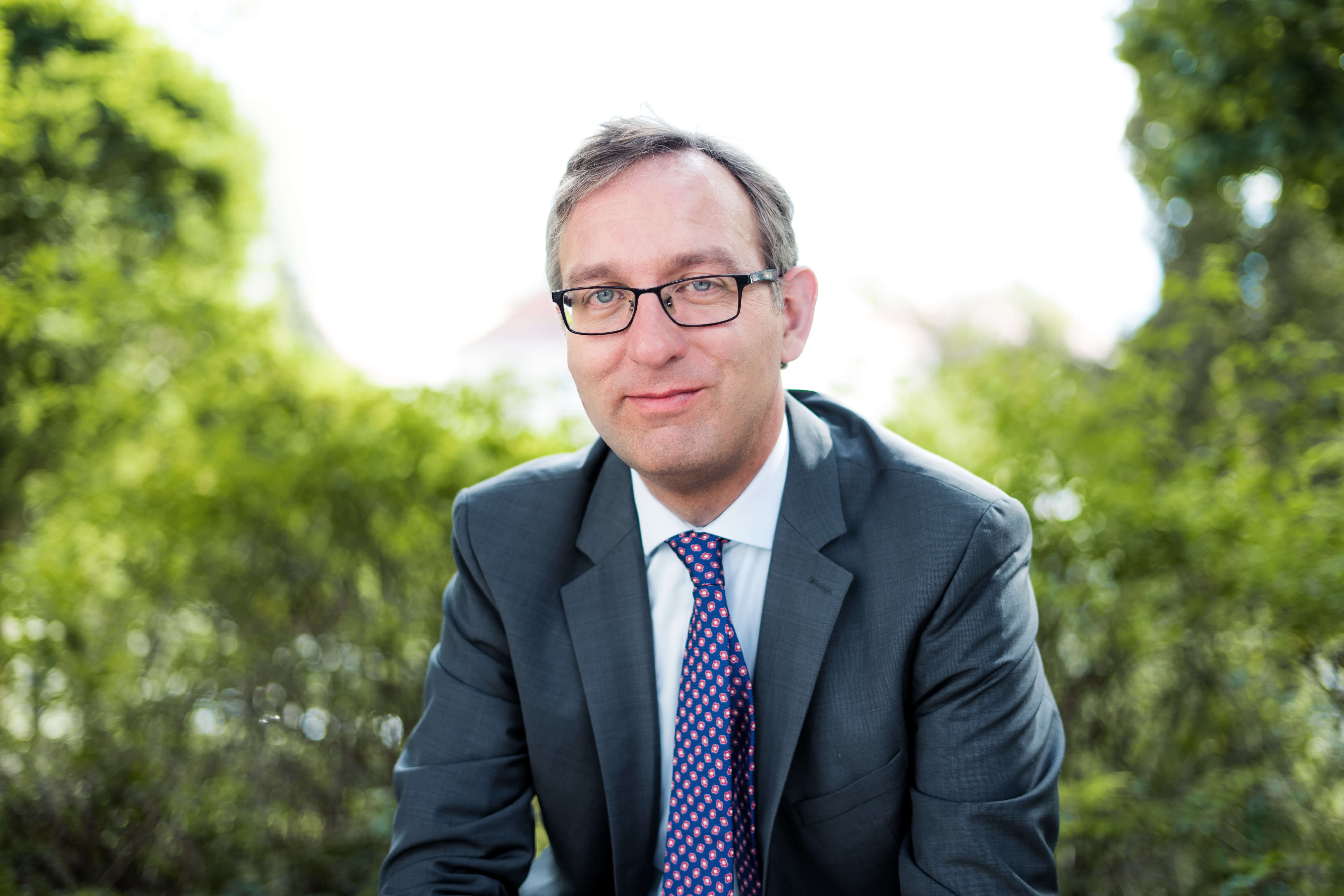 Ein lächelnder Mann im Anzug mit Brille und Krawatte sitzt vor grünem, unscharfem Hintergrund.