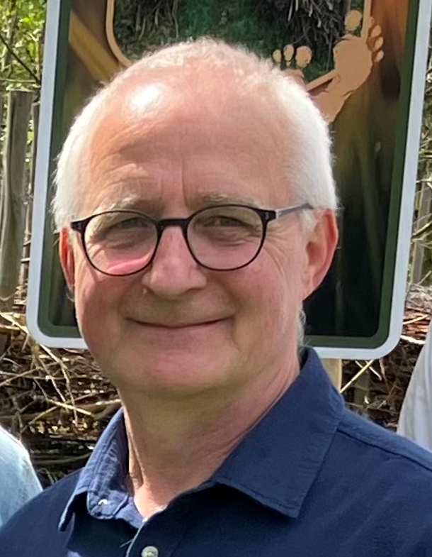 Ein lächelnder Mann mit Brille und weißen Haaren trägt ein dunkelblaues Hemd vor einem Schild.