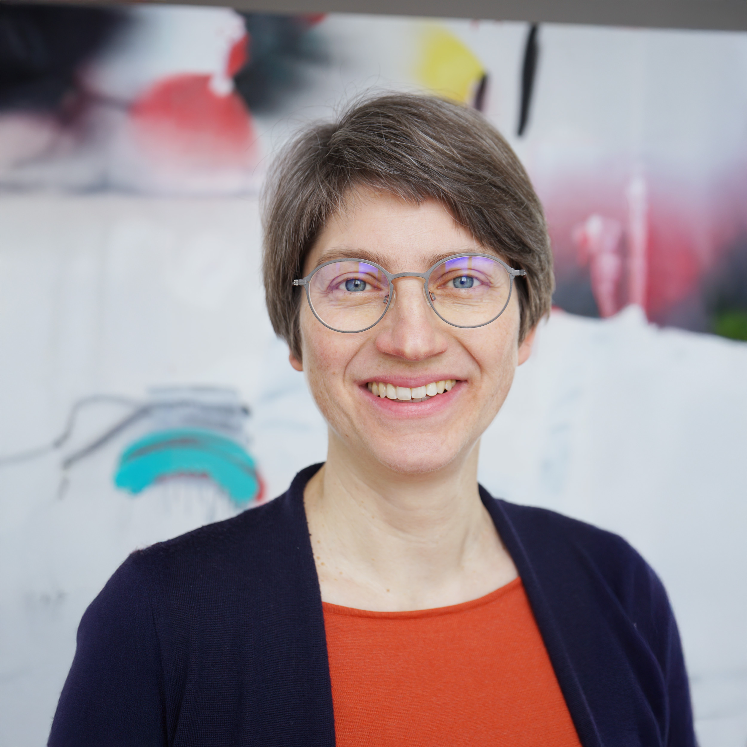 Eine lächelnde Frau mit kurzen Haaren und runder Brille vor einem hellen, farbigen Hintergrund.