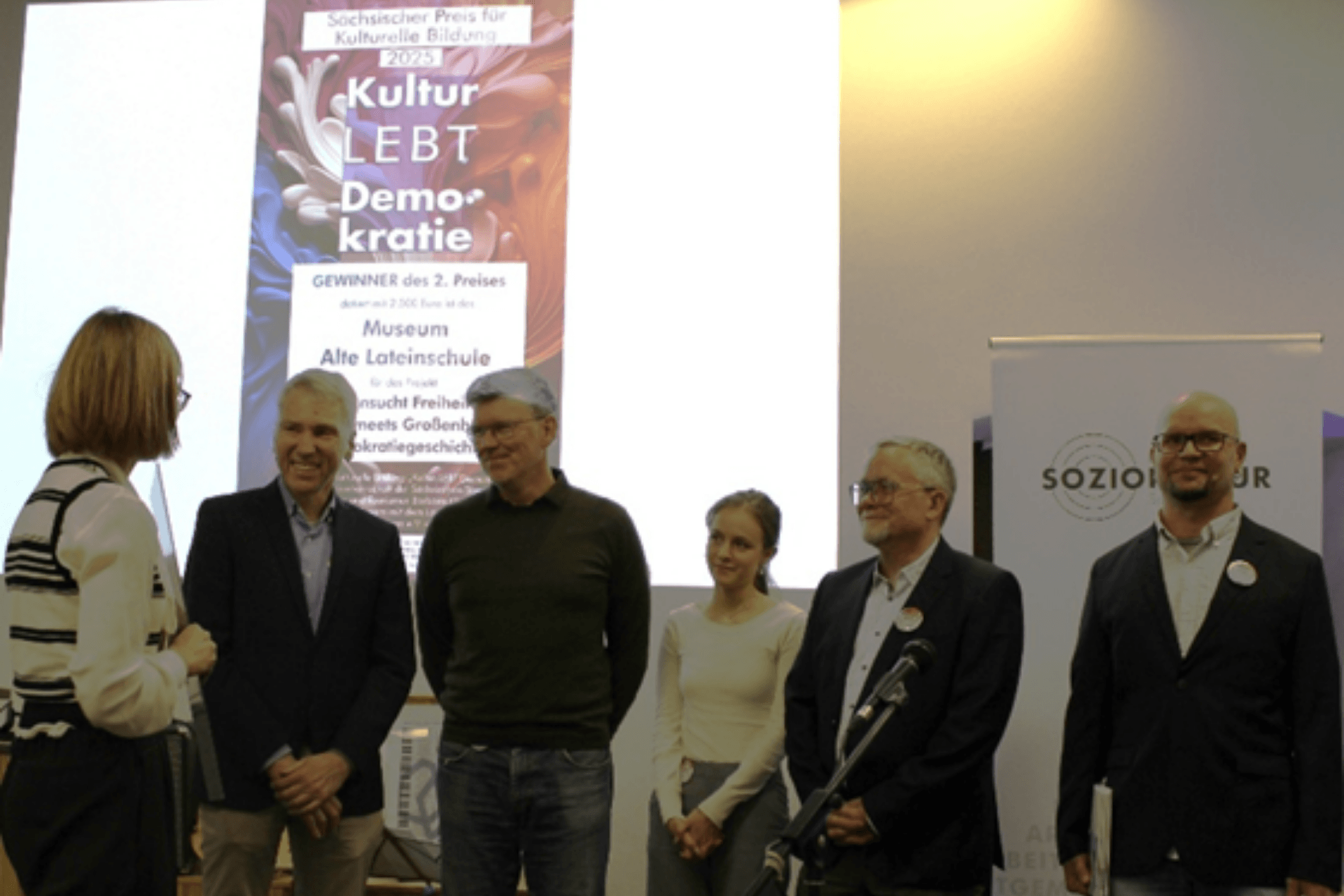 Eine Preisverleihung auf einer Bühne. Fünf Personen stehen nebeneinander vor einer großen Leinwand, auf der ein Plakat mit dem Titel „Sächsischer Preis für Kulturelle Bildung 2024 – Kultur LEBT Demokratie“ zu sehen ist. Der Text auf der Leinwand verkündet das „Museum Alte Lateinschule“ als Gewinner des 2. Preises. Die Personen – vier Männer in geschäftlicher Kleidung und eine junge Frau in einem hellen Oberteil – blicken lächelnd zu einer Frau am linken Bildrand, die ihnen den Preis überreicht. Rechts im Bild steht ein Roll-up-Banner mit dem Logo „Soziokultur“. Die Szene wirkt feierlich und wertschätzend.