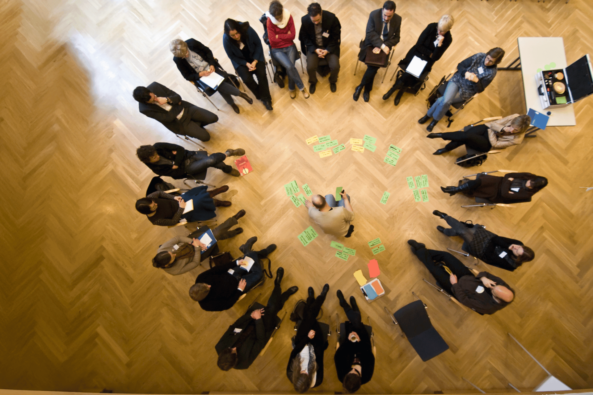 Eine Luftaufnahme einer Gruppe von 16 Personen, die in einem weiten Stuhlkreis auf einem hellen Holzparkettboden sitzen. In der Mitte des Kreises kniet eine Person auf dem Boden und ordnet zahlreiche grüne und gelbe Moderationskarten an, die sternförmig um sie herum verteilt sind. Die Teilnehmer, überwiegend in Business-Casual gekleidet, beobachten den Prozess aufmerksam, einige machen sich Notizen. Die Szene wirkt wie ein intensiver Workshop oder eine Brainstorming-Session in einem großzügigen, lichtdurchfluteten Saal.