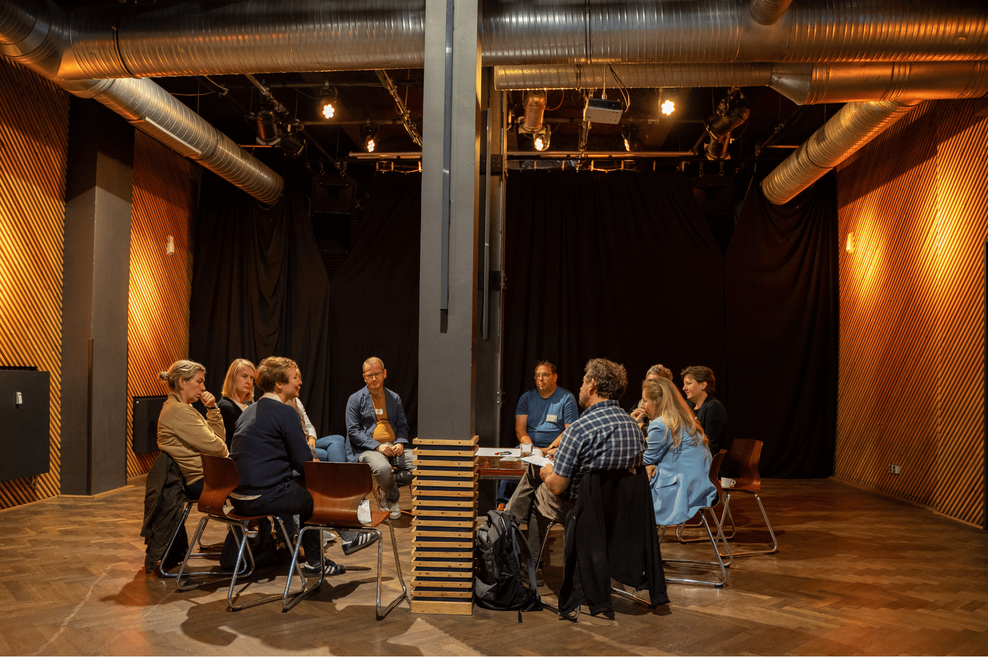 Eine Gruppe von neun Personen sitzt in einem großen, modernen Veranstaltungsraum im Kreis zusammen. Der Raum besticht durch ein industrielles Design mit sichtbaren Lüftungsrohren an der Decke, dunklen Vorhängen im Hintergrund und Wänden mit einer markanten, gelb-gestreiften Holzoptik. In der Mitte des Kreises steht eine tragende Säule, die teilweise mit Holzlamellen verkleidet ist. Die Teilnehmenden sitzen auf schlichten Holzstühlen und sind in ein Gespräch vertieft, wobei eine Person im blauen Sakko und eine weitere im karierten Hemd besonders präsent wirken. Die warme Beleuchtung sorgt für eine konzentrierte Arbeitsatmosphäre.