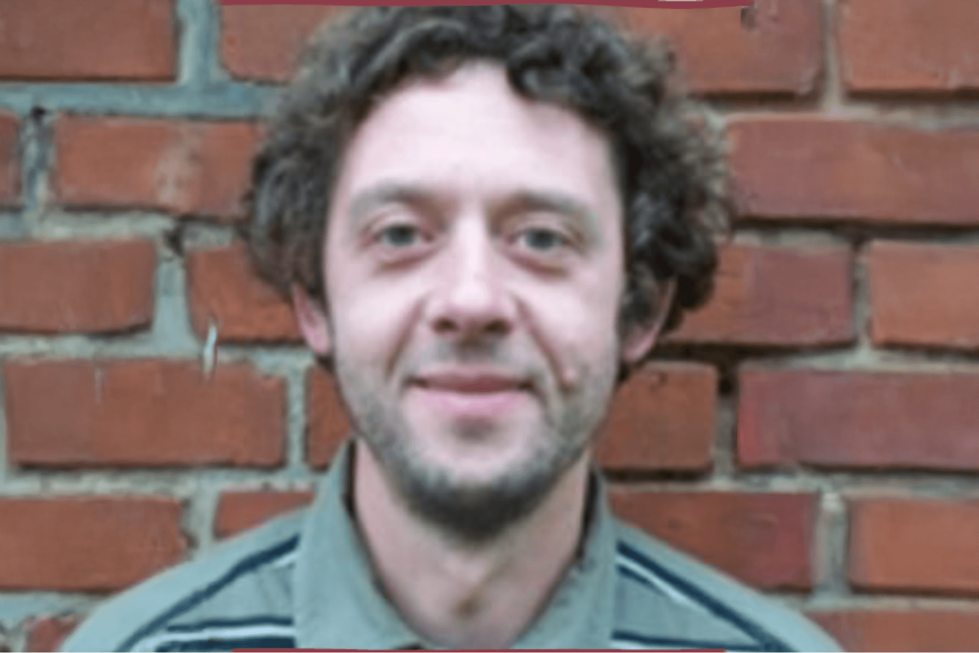Ein Porträtfoto eines lächelnden Mannes mit lockigem, braunem Haar und einem kurzen Bart. Er trägt ein grünliches Hemd mit dunklen Streifen am Kragen. Der Hintergrund besteht aus einer rustikalen roten Backsteinwand. Der Bildausschnitt ist eng auf das Gesicht und die Schultern fokussiert.