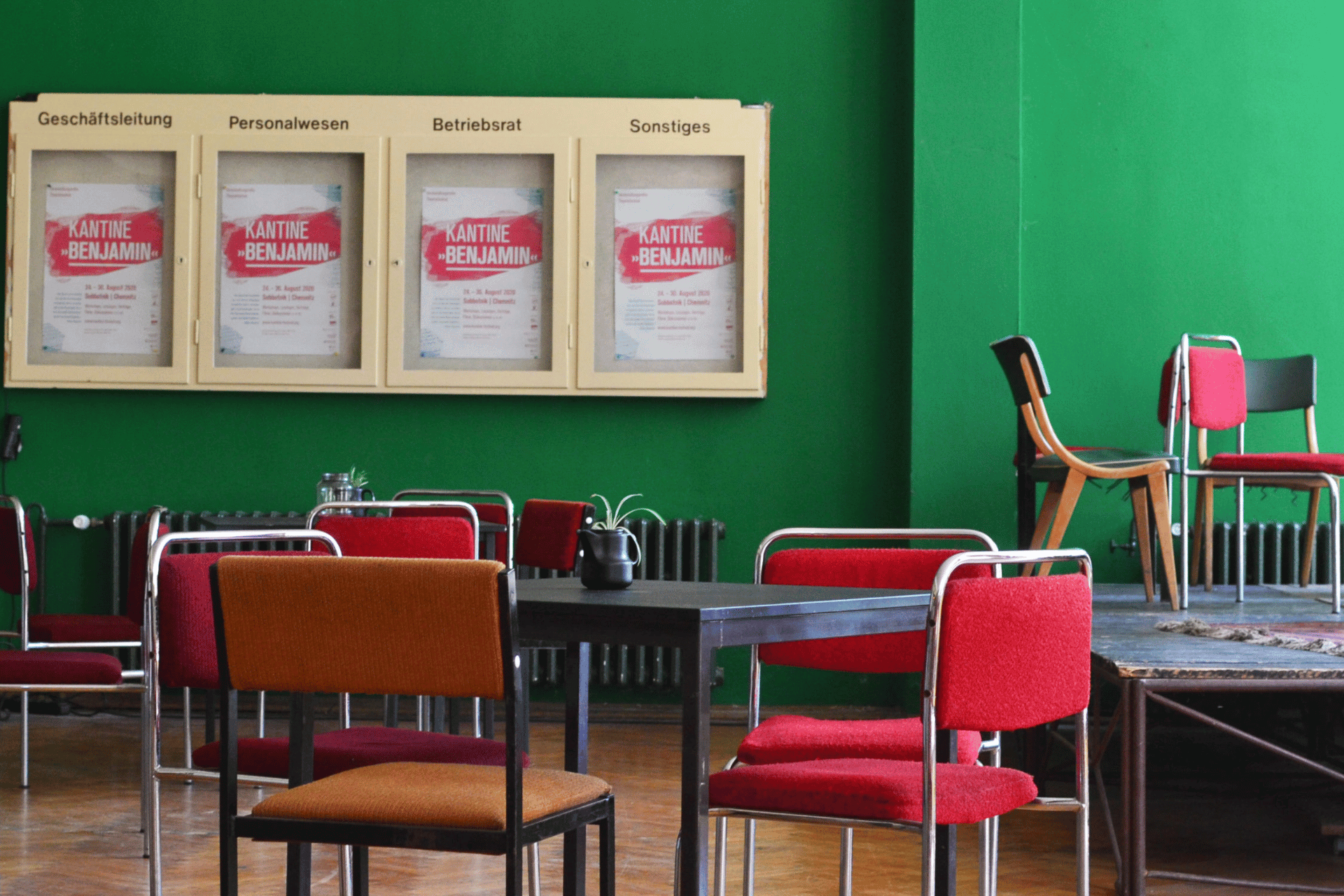 Ein Innenraum mit einer auffällig leuchtend grünen Wand. An der Wand hängt ein heller Schaukasten mit vier Glassegmenten, die mit den Beschriftungen „Geschäftsleitung“, „Personalwesen“, „Betriebsrat“ und „Sonstiges“ versehen sind. In jedem Segment hängt das gleiche rot-weiße Plakat mit dem Titel „KANTINE BENJAMIN“. Im Vordergrund stehen mehrere schwarze Tische mit roten und orangefarbenen Polsterstühlen auf einem Parkettboden. Rechts im Bild ist eine leicht erhöhte Holzbühne erkennbar, auf der ebenfalls Stühle stehen. Die Szene wirkt wie ein retro-charmanter Aufenthaltsraum oder ein Kulturzentrum.