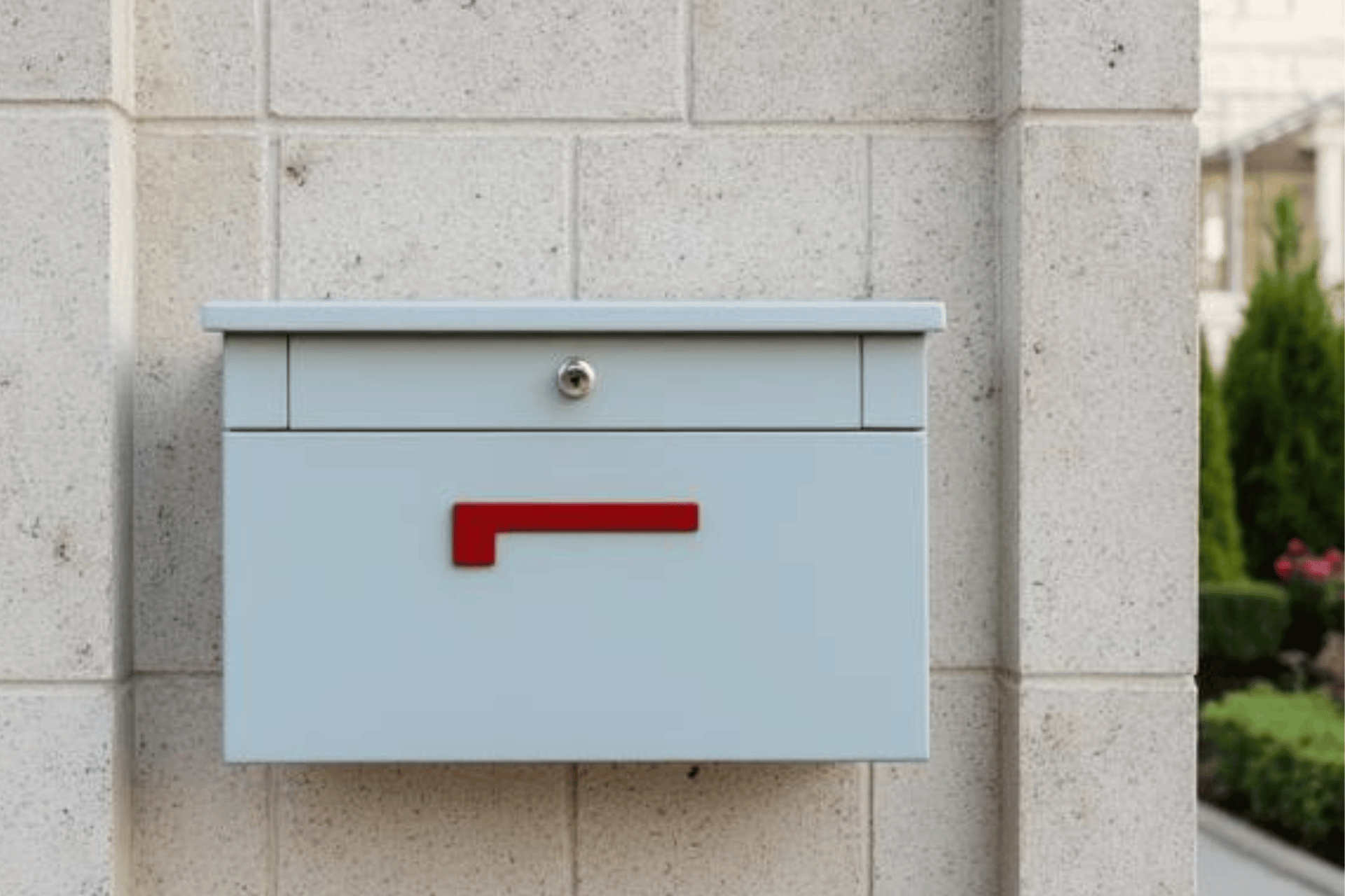 Ein moderner, hellgrauer Briefkasten, der an einer Wand aus hellen, rechteckigen Steinen montiert ist. Der Briefkasten hat ein schlichtes, minimalistisches Design mit einem kleinen Schlüsselschloss an der oberen Klappe. Auffällig ist ein rotes, rechtwinkliges Element (ein Griff oder eine Markierung) in der Mitte der Frontseite. Im Hintergrund ist rechts ein kleiner Teil einer gepflegten Außenanlage mit grünen Hecken und Pflanzen zu erkennen.
