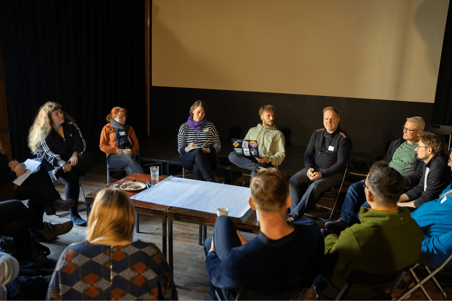 Eine Gruppe von etwa zehn Personen sitzt in einem abgedunkelten Raum in einem Stuhlkreis zusammen. In der Mitte steht ein niedriger Holztisch, auf dem ein großer weißer Papierbogen, ein Glas Wasser und eine kleine Schale liegen. Die Teilnehmenden, überwiegend in Alltagskleidung, sind in ein Gespräch vertieft. Ein Mann in der Mitte hält einen Laptop mit vielen Aufklebern auf dem Schoß, während eine Frau daneben Notizen in ein Buch schreibt. Im Hintergrund befindet sich eine große, helle Leinwand und ein schwarzer Vorhang, was auf einen Seminar- oder Kinoraum hindeutet.