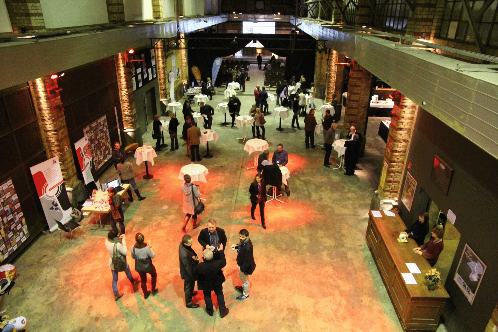 Eine Weitwinkel-Aufnahme aus erhöhter Perspektive von einem Empfang oder einer Netzwerk-Veranstaltung in einer großen Industriehalle mit unverputzten Backsteinwänden. Zahlreiche Personen stehen in kleinen Gruppen an weißen Stehtischen verteilt im Raum und unterhalten sich. Der Boden ist durch rötliche Scheinwerfer punktuell beleuchtet. Am linken Bildrand befinden sich Informationstische und Roll-up-Banner, während auf der rechten Seite ein hölzerner Empfangstresen zu sehen ist. Im Hintergrund führt der Blick in einen weiteren, dunkleren Bereich der Halle, in dem Stuhlreihen erkennbar sind.